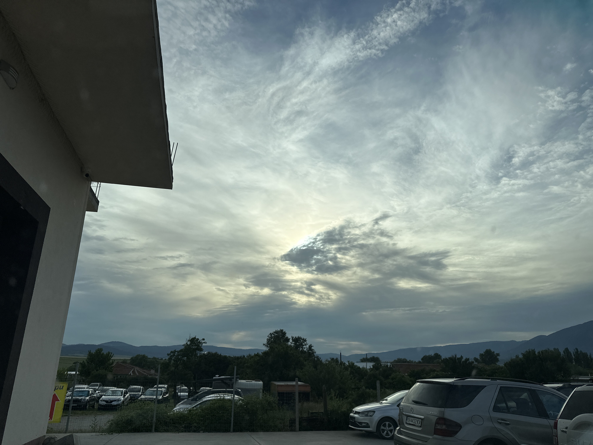 In Piperevo, Bulgaria, on a cloudy day with the sun partially obscured by streaks of cirrus clouds, an outdoor parking lot is visible. A white building with a dark overhang and a security camera is in the foreground on the left. The parking lot contains numerous cars, including a silver SUV with the license plate "YH 4432 KA" on the right. Beyond the parking lot, a treeline is present, and in the distance, rolling hills or mountains can be seen under the overcast sky.