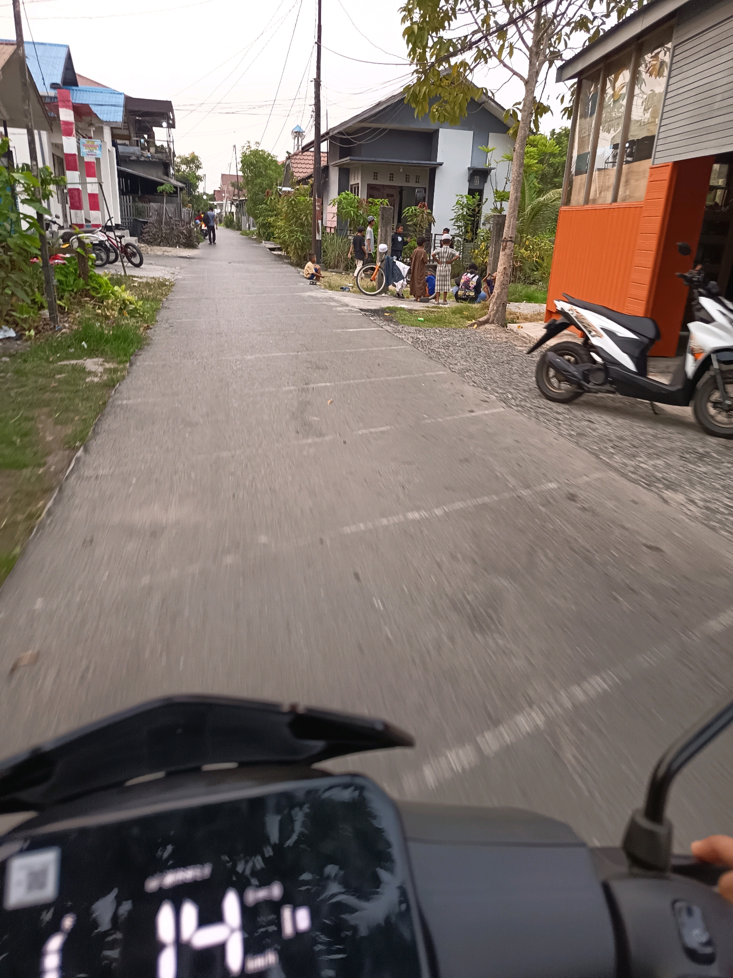 The image depicts a street scene in Banjarmasin, Indonesia, as viewed from the perspective of someone riding a motorcycle. The street is paved and marked with white lines, suggesting it may be a parking area or a road within a residential or commercial district. On the left side of the street, there are houses with red and white striped columns and blue roofs, interspersed with greenery. On the right side, a building with an orange base and large glass windows is visible, with a white and black scooter parked in front of it.
Several people are present in the scene, appearing to be a mix of adults and children. Some are standing near a house on the right, while others are closer to the street. One child is sitting on the ground near a bicycle. The overall atmosphere suggests a casual daytime activity. The motorcycle from which the photo is taken has a digital display showing a speed of "14". The sky is overcast.