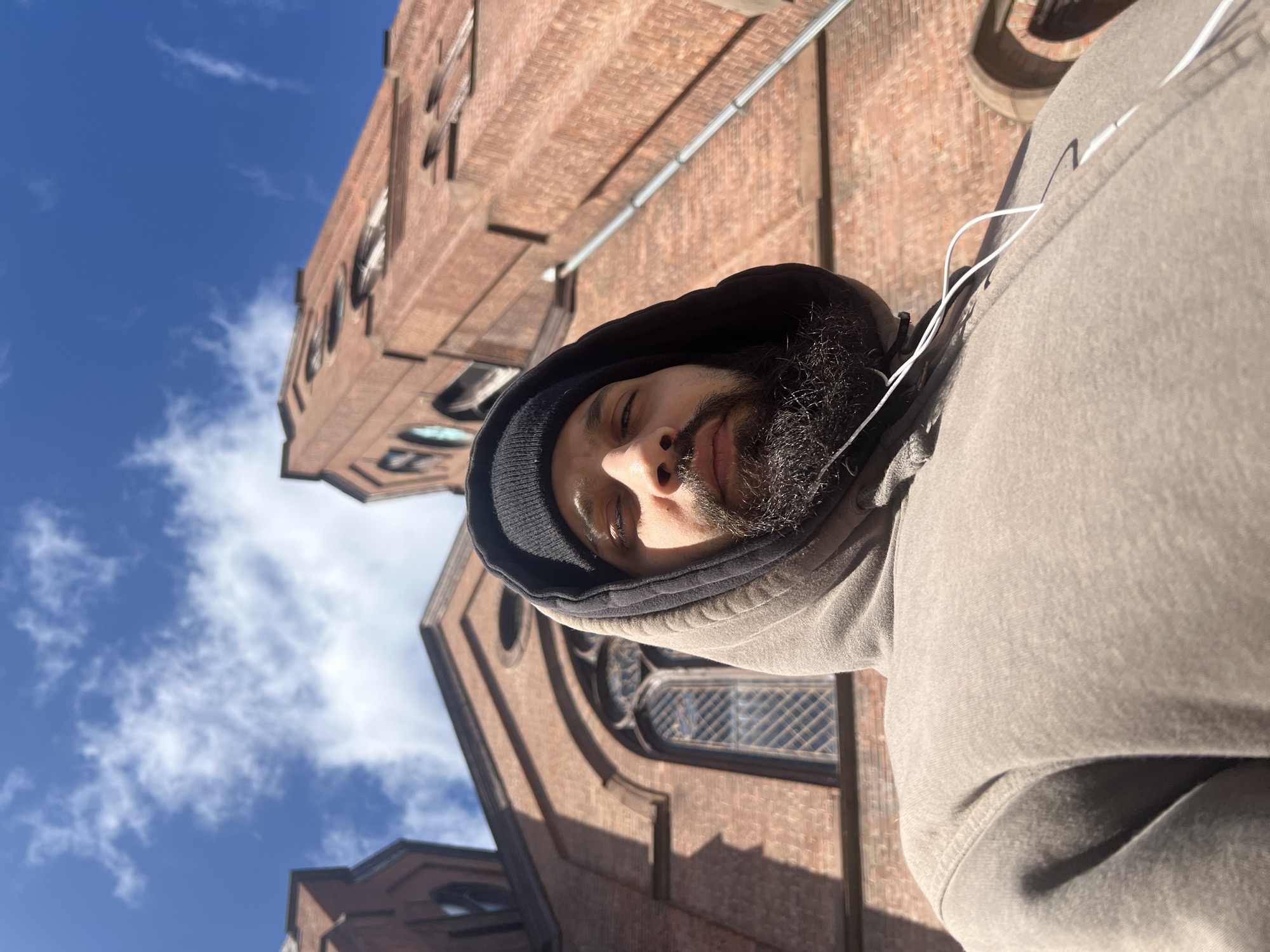 A low-angle, outdoor shot of a man with a beard and mustache. He is wearing a gray hooded sweatshirt, with the hood up and a black beanie underneath. White earphones are visible near his neck. The background is a brick building with arched windows. The sky is visible in the upper-left corner, with some clouds. The perspective suggests that the man is looking down at the camera. The photograph appears to be taken in the City of Newburgh, United States of America, given the context.