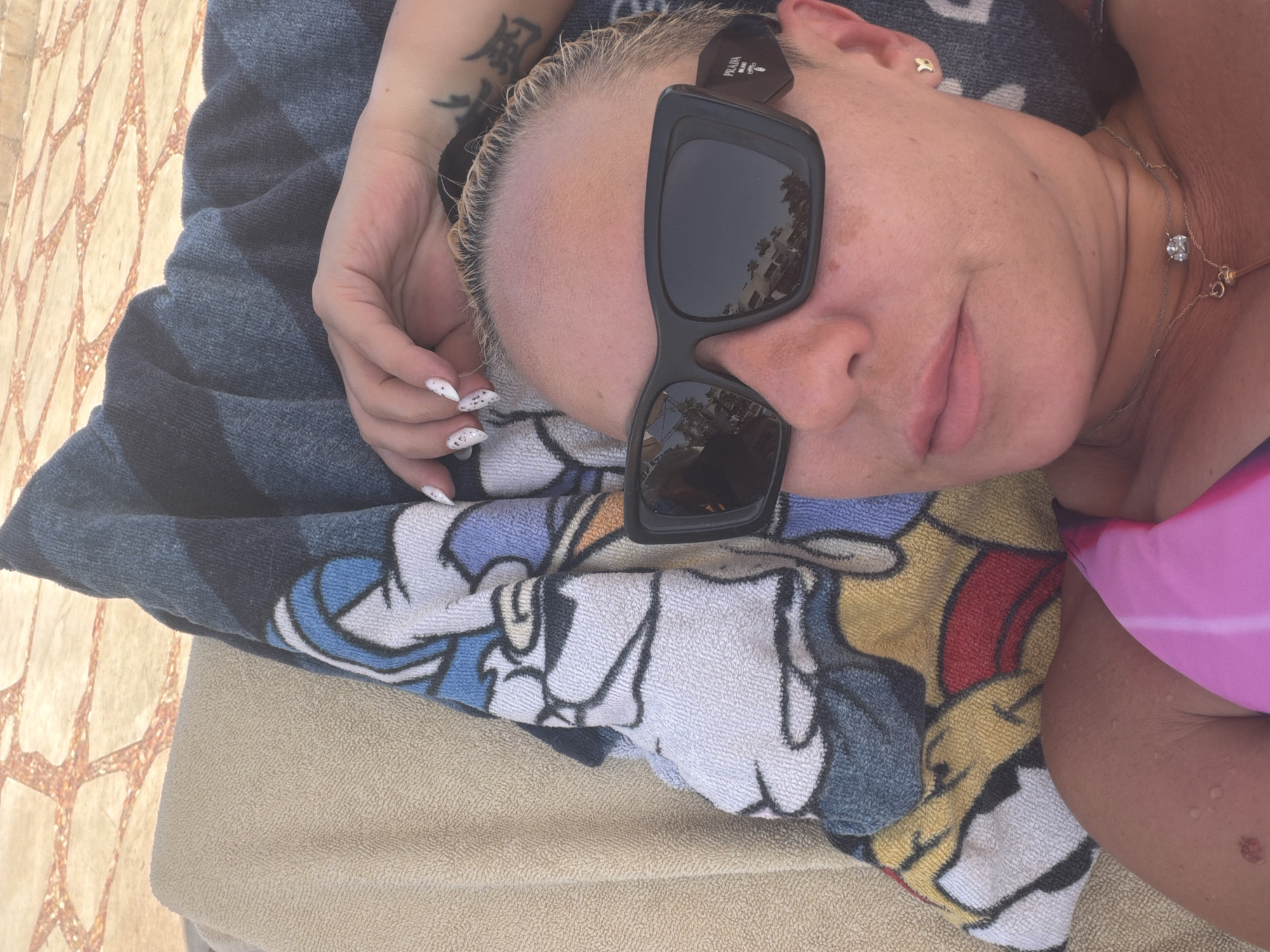 A woman with blonde hair and sunglasses is lying on a lounge chair, covered by a towel with cartoon characters. The scene is outdoors, likely at a resort in Sharm El Sheikh, Egypt, as indicated by the tiled patio visible in the background. The woman is wearing a pink bikini top and a silver necklace. Her hand is resting on the towel, and she has a tattoo on her arm. The lighting suggests it is daytime.