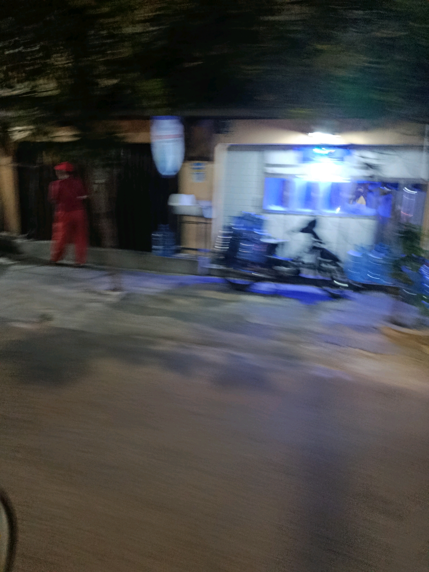 A blurry, nighttime photograph taken in front of a building in Bekasi, Indonesia, shows a person in red clothing standing on the left. In the background, a white building has bright blue lights illuminating its interior. A motorcycle is parked in front of the building, with several large water jugs nearby. The ground is wet, reflecting the lights. The overall impression is of a dimly lit, urban scene.