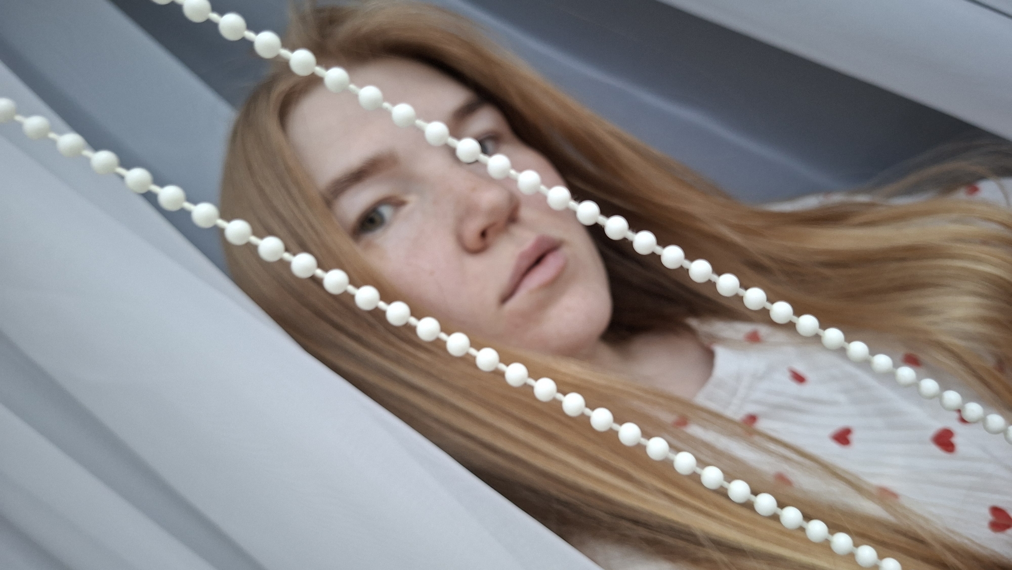 A young, light-skinned woman with reddish-blonde hair is pictured indoors. She is wearing a light-colored top with red heart-shaped patterns. The woman's face is partially obscured by a white beaded pull chain that runs diagonally across the foreground of the image. The background appears to be a window with closed white blinds. The setting might be a residential area in Chaikovsky, Russia.