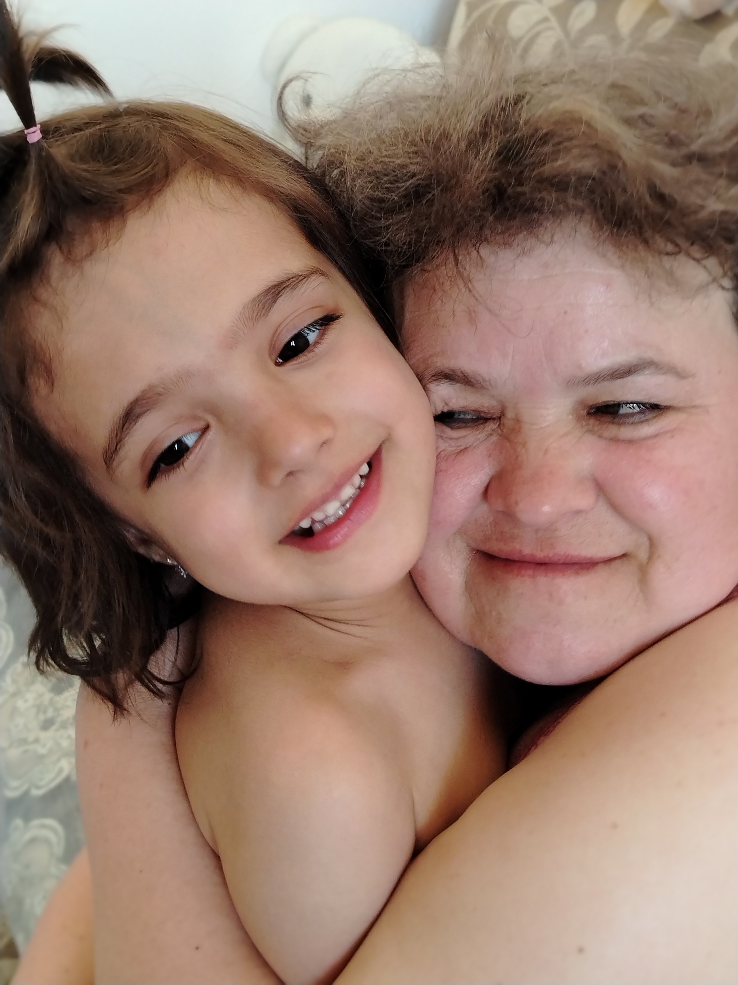 The image features a close-up of a child and an adult, both appearing to be female. The child, likely a girl, is younger with brown hair pulled back into a small ponytail secured with a pink hair tie. She is smiling and hugging the adult. The adult has curly hair and is also smiling, with their faces close together. The background is out of focus, suggesting a domestic setting. There is no discernible information to indicate a location in the city of Noviy, Russia. There are no other objects or environmental details that stand out. The interaction suggests a familial or affectionate relationship.