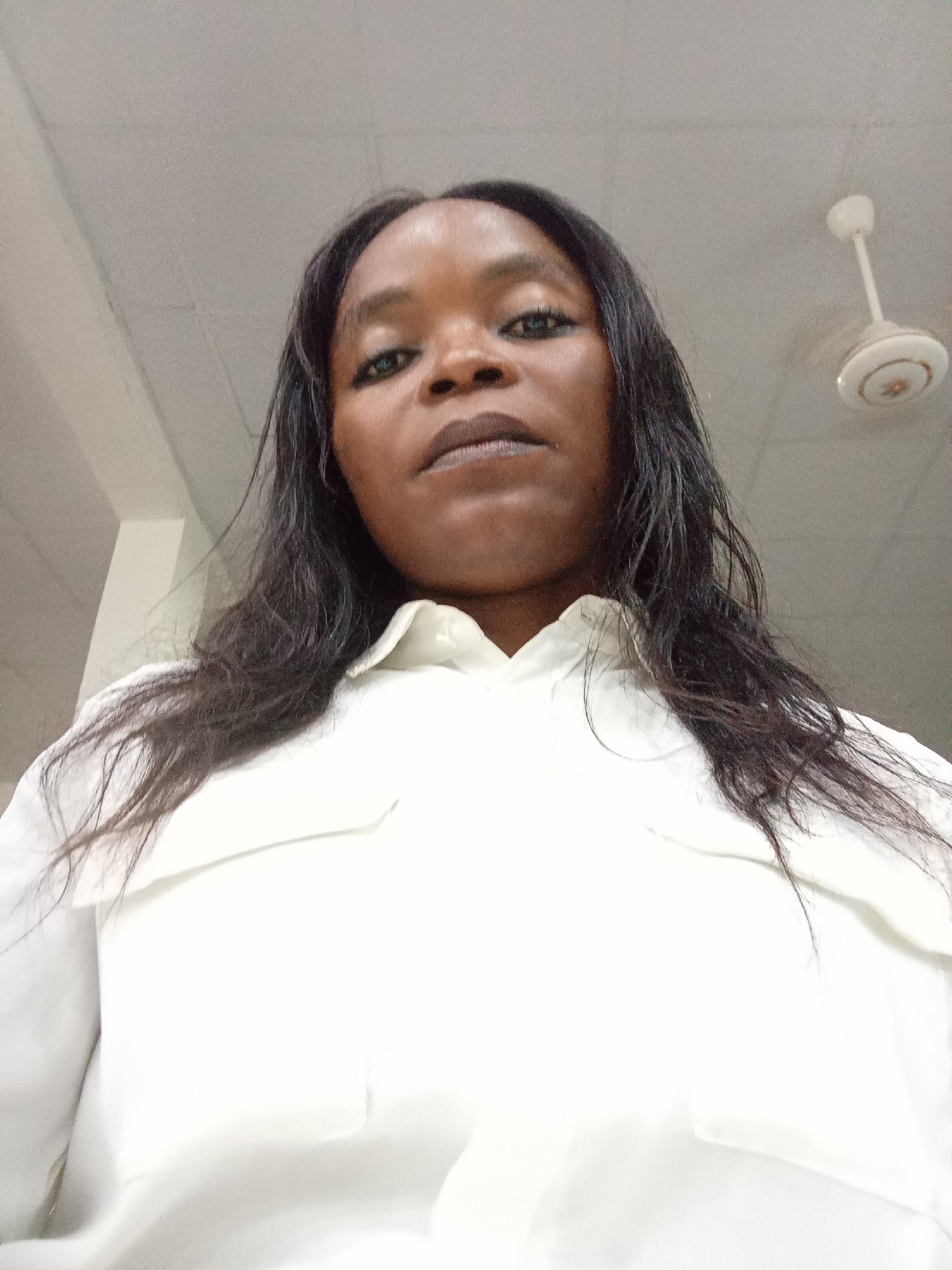 A low-angle, medium shot shows a Black woman from the chest up, looking directly into the camera. She has long, dark, slightly wavy hair that falls past her shoulders and is wearing a white collared shirt. Her makeup includes dark eyeliner and mascara, and her lips are painted a neutral color. The background appears to be an indoor space with a white, tiled ceiling and a white ceiling fan with gold accents visible in the upper right corner. The lighting is bright and even. No specific location within Kason, Nigeria, can be identified from the image.
