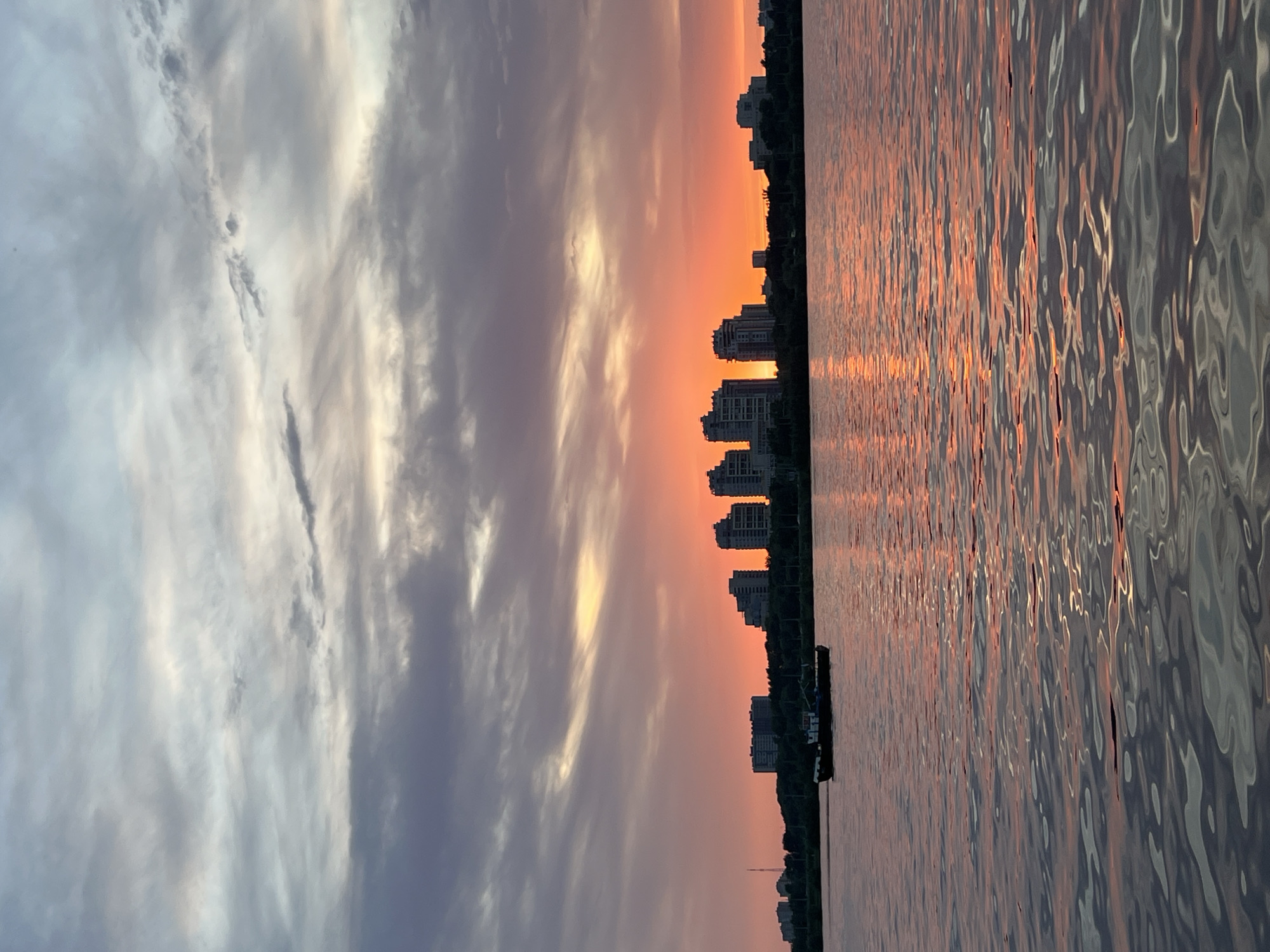 The image shows a sunset over water in Kazan, Russia. The sky is filled with clouds in shades of gray, white, orange, and pink. The sun is setting behind a row of buildings along the shoreline. The water is reflecting the colors of the sky, with a shimmering effect. There is a small boat anchored near the shoreline. The buildings appear to be residential.