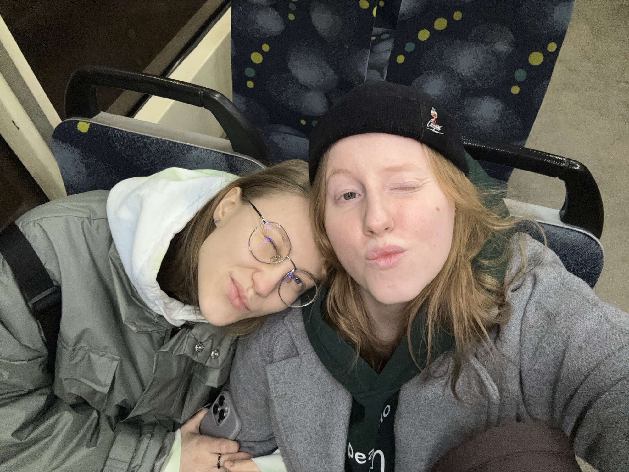 Two young women are posing for a selfie while seated on public transport. The woman on the left has brown hair, is wearing round glasses, and has her head resting on the other woman's shoulder. She is wearing a grey hooded coat and is holding a phone in her left hand. The woman on the right is wearing a black beanie with a logo on it and winking. She is wearing a grey coat over a green hoodie. The seats of the public transport have a dark blue patterned upholstery with yellow and green dots. The photo appears to have been taken indoors, likely in a bus or train in Perm, Russia, evidenced by the distinctive seat upholstery commonly found in public transport within the city.