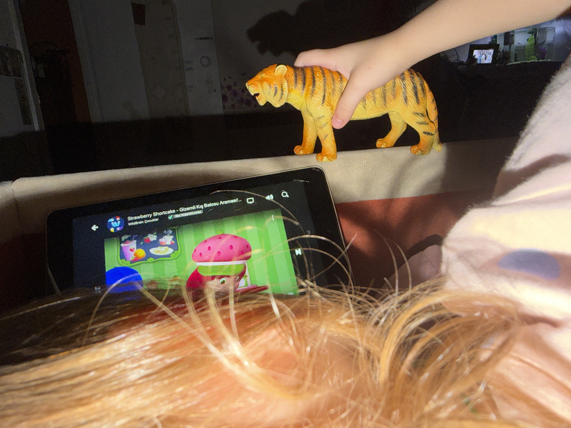 In Dnipro, Ukraine, a child's hand is holding a toy tiger, positioned above a tablet screen displaying an episode of "Strawberry Shortcake". The tablet is lying on a textured surface, possibly a couch, with the head and blonde hair of a person in the foreground. The background is dimly lit, with hints of furniture and a distant television screen showing a small image, possibly another toy or a decorative item. The lighting suggests an indoor setting, likely a home environment.