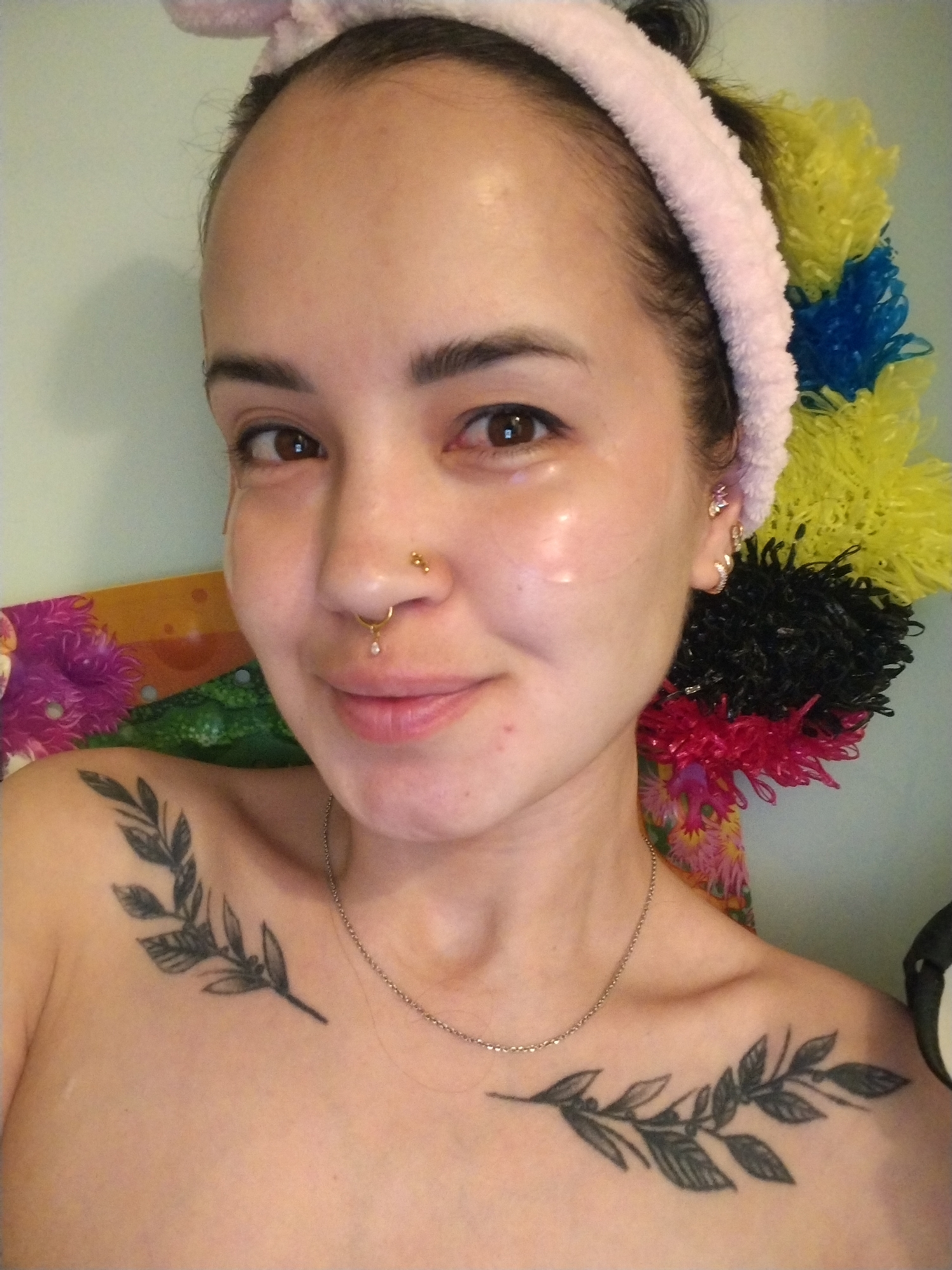 A woman with light skin is centered in the image, wearing a pink headband and a silver necklace. She has eyebrow piercings and a nose ring, and her face has a glossy sheen. She has tattoos of leafy branches on her chest. Behind her is a colorful, feathery backdrop. The environment appears to be indoors with a light green wall.