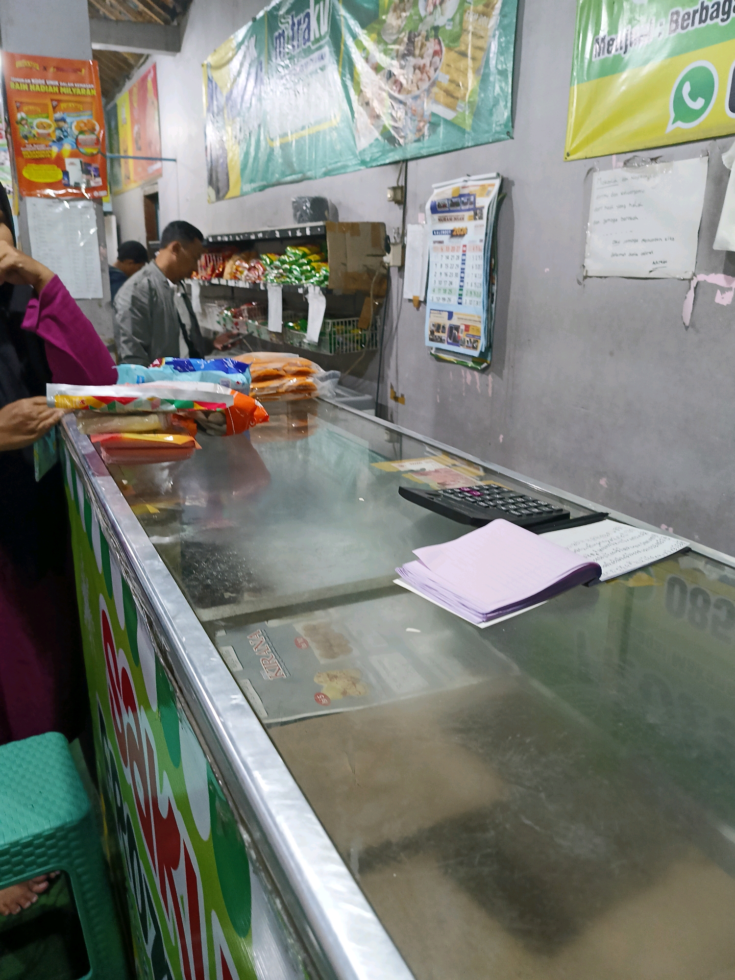 The image depicts the interior of a small shop in Manisrenggo, Indonesia, likely a minimarket or convenience store. A glass-top freezer display case dominates the foreground, filled with various packaged food items. On top of the freezer, a black calculator and a stack of purple paper are visible. Behind the freezer, a man in a grey jacket is seen interacting with the shopkeeper or another customer, standing in front of shelves stocked with more merchandise. Banners and posters are displayed on the wall, including a calendar and advertisements. The overall environment appears to be a typical retail setting with a focus on food products.