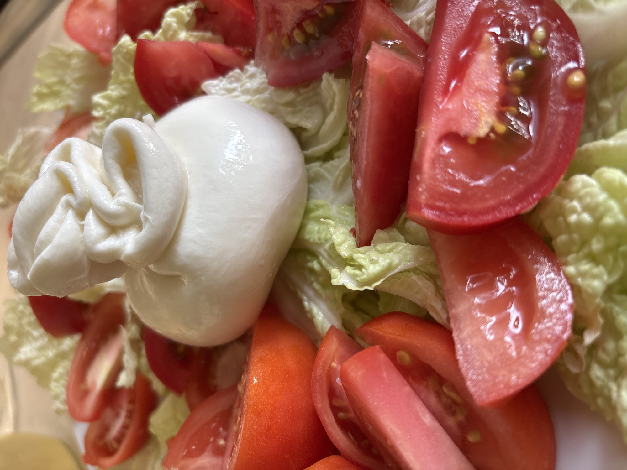 A close-up, overhead shot showcases a fresh salad centered on a white plate. The salad is composed of vibrant red tomato wedges arranged around a plump ball of fresh mozzarella cheese. The mozzarella, with its characteristic twisted top, sits atop a bed of shredded lettuce. The scene is bathed in natural light, highlighting the textures and colors of the ingredients. There is no indication of a specific location in Rusanovo, Russia, or any people present.