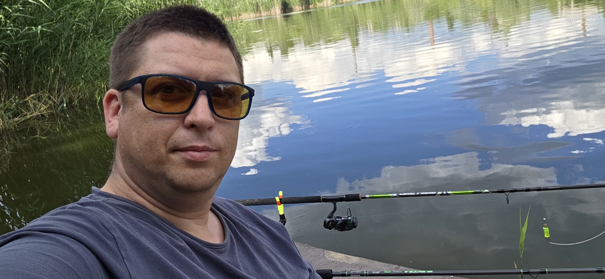 This is a selfie of a man wearing sunglasses with orange lenses, a dark grey t-shirt, and a short haircut. He is positioned on the left side of the frame, looking towards the camera.
The background depicts a calm body of water with reflections of the sky, which appears to be blue with white clouds. Lush green reeds or tall grass grow along the left bank of the water. In the foreground, two fishing rods are visible, resting horizontally and extending across the lower portion of the image. One rod has a fishing reel attached and a yellow and orange bobber. The other rod, positioned lower, also has a green design and a yellow bobber.
The scene suggests the man is engaged in or about to engage in fishing. The lighting indicates it is daytime, likely during the day, with a bright sky. The overall atmosphere appears relaxed and casual, consistent with an outdoor recreational activity. There is no visible text in the image.