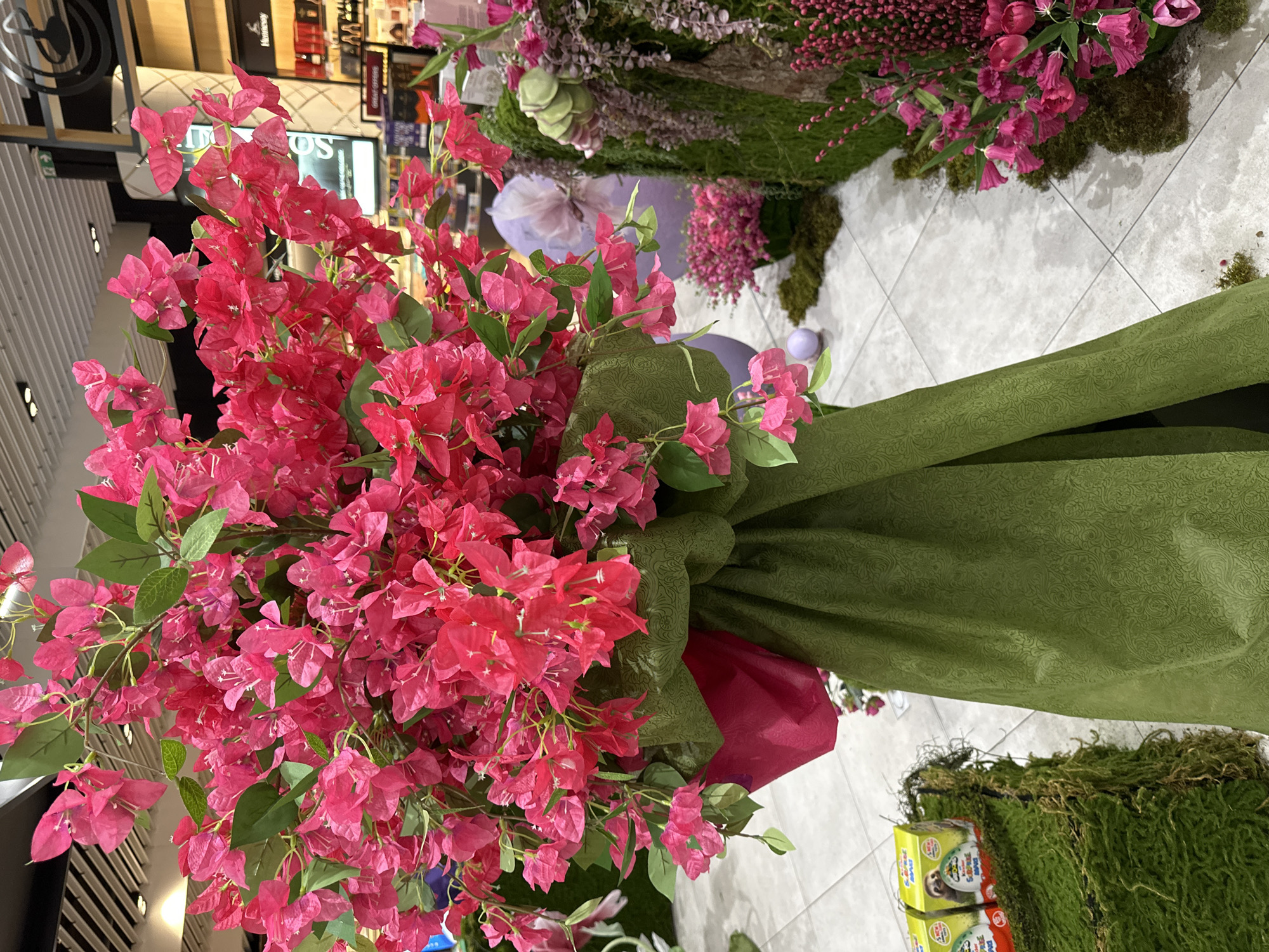 The image depicts a large bouquet of pink bougainvillea flowers wrapped in green and red paper, likely displayed for sale or decoration. The bouquet sits on a light-colored tile floor. Behind the bouquet, there is an arrangement of pink and purple flowers and moss-covered structures. In the background, shelves with various items suggest this is likely a store or shop interior, possibly in Sofia, Bulgaria, though specific clues for the city are absent in the image. There are no people visible in the immediate foreground.
