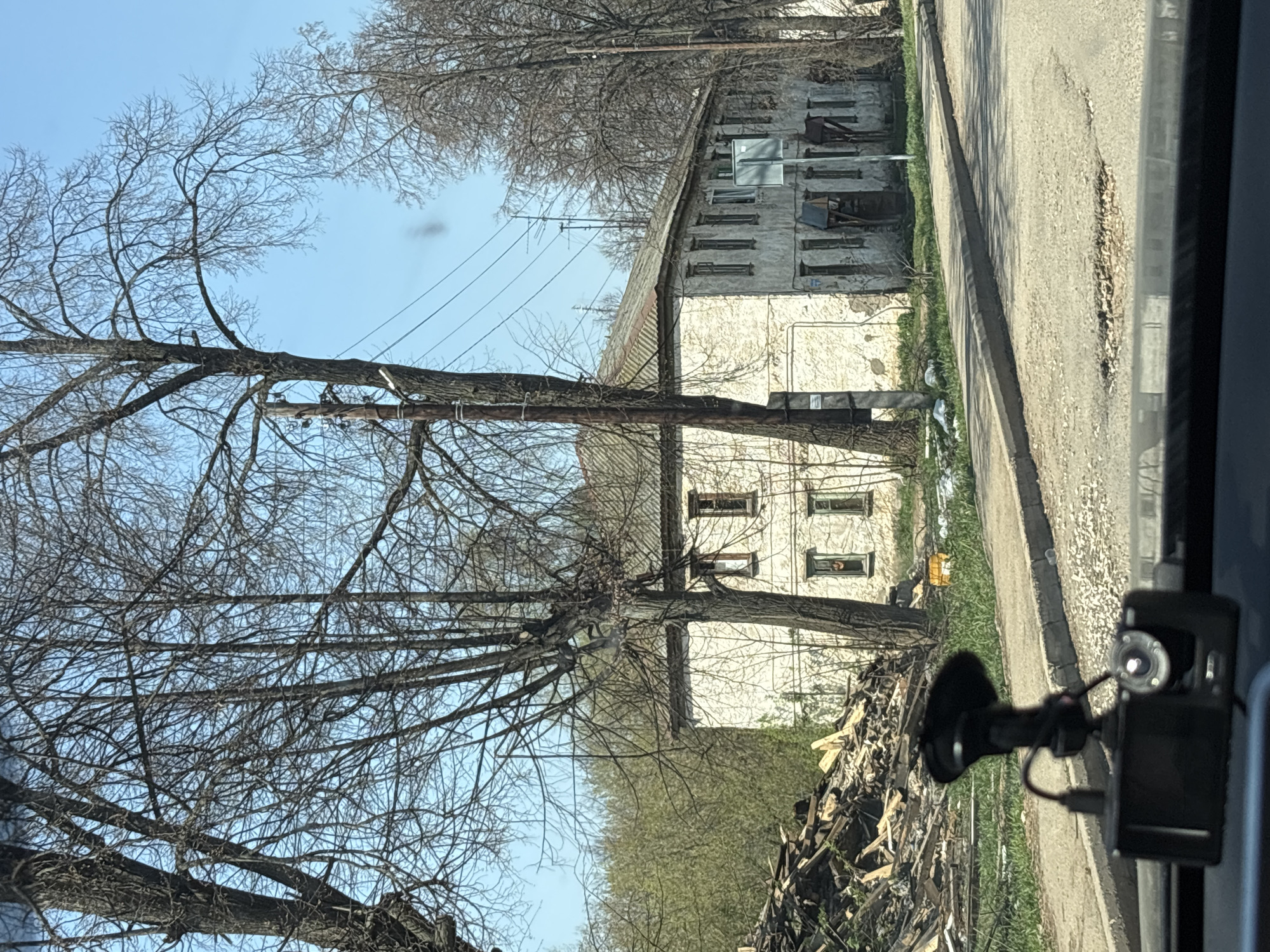 The image depicts an old, white, multi-story building with numerous windows, some of which are boarded up or damaged. The building appears dilapidated and possibly abandoned. In front of the building, a large pile of debris and rubble is visible. A utility pole stands in the foreground, partially obscured by bare tree branches. The sky is light blue. The image seems to have been taken from a vehicle traveling along a paved road that runs alongside the building. A car dashboard camera is visible in the lower right corner. Given the context provided, the location is presumed to be within the city of Chaikovsky, Russia.