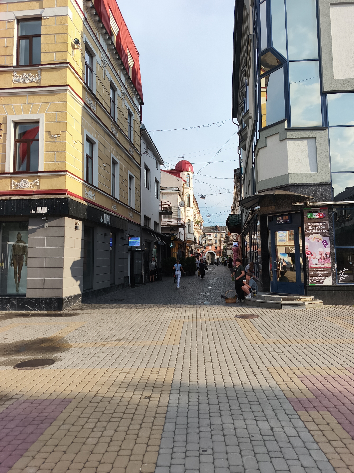 A sunlit cobblestone street is flanked by two buildings. The building on the left is a vintage yellow structure with red trim and white ornate details. It features several windows and a shop on the ground floor with a mannequin displayed in the window. The building on the right is modern with a glass facade reflecting the sky and clouds. It has a dark blue entrance with signs and advertisements. People are seen walking and sitting on steps in the distance. The street is paved with interlocking pavers in shades of yellow, gray, and pink. A manhole cover is visible in the foreground pavement. The scene appears to be in a European city, potentially Ternopil, Ukraine, given the architecture and signage.