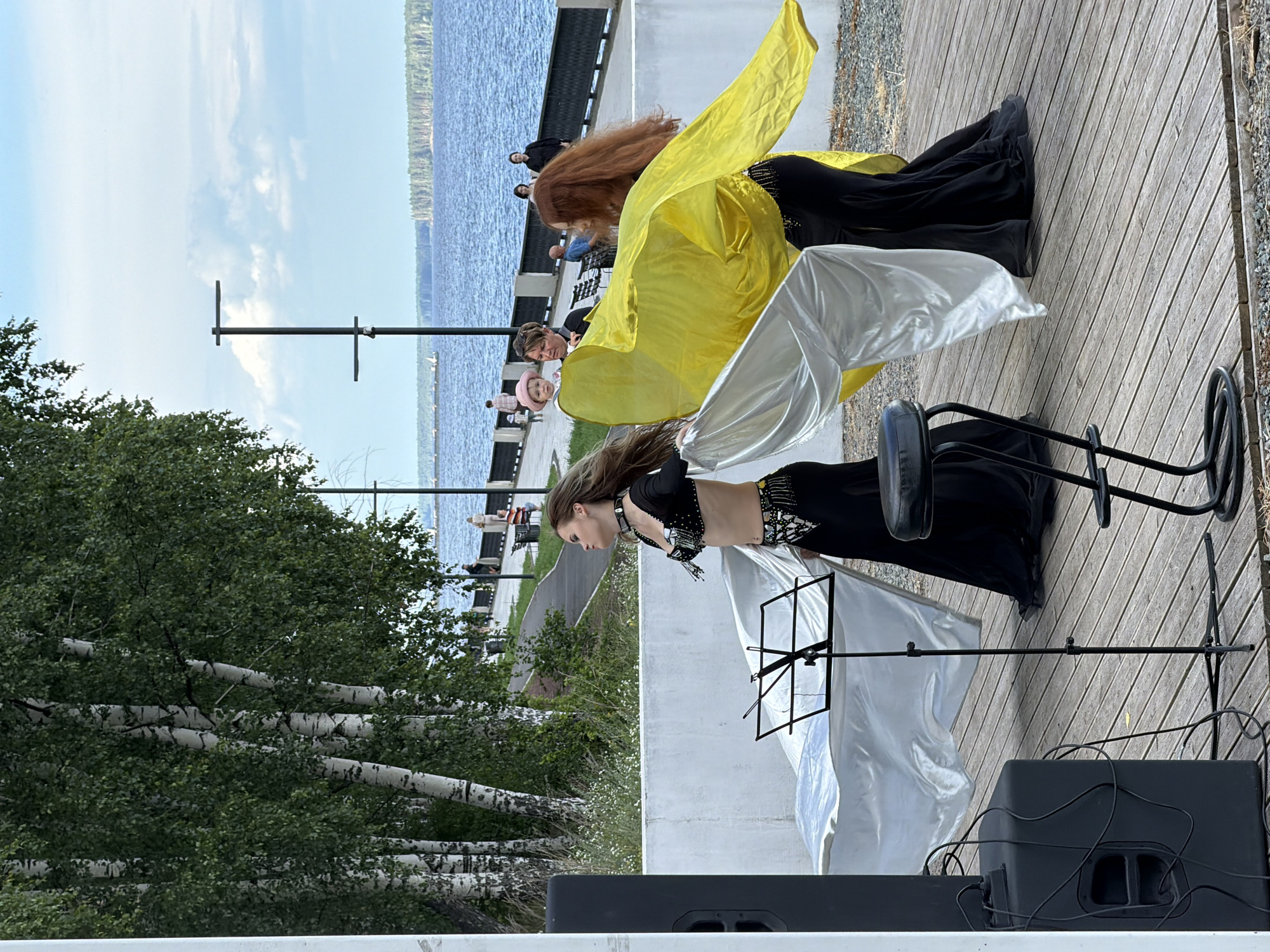 In Chaikovsky, Russia, a daytime outdoor event is underway near a large body of water, likely the Kama River. Two female belly dancers are performing on a wooden boardwalk. The dancer on the left, wearing a black costume with beaded embellishments, holds a large white fabric. The dancer on the right, also in a black costume, holds a large yellow fabric. Behind them, a white concrete wall separates the boardwalk from a grassy area with birch trees. Further back, people are walking along a paved pathway next to a dark metal fence. The background features the river and a distant, treed shoreline under a partly cloudy sky. A black speaker is visible to the left of the frame, and a music stand is placed between the dancers.