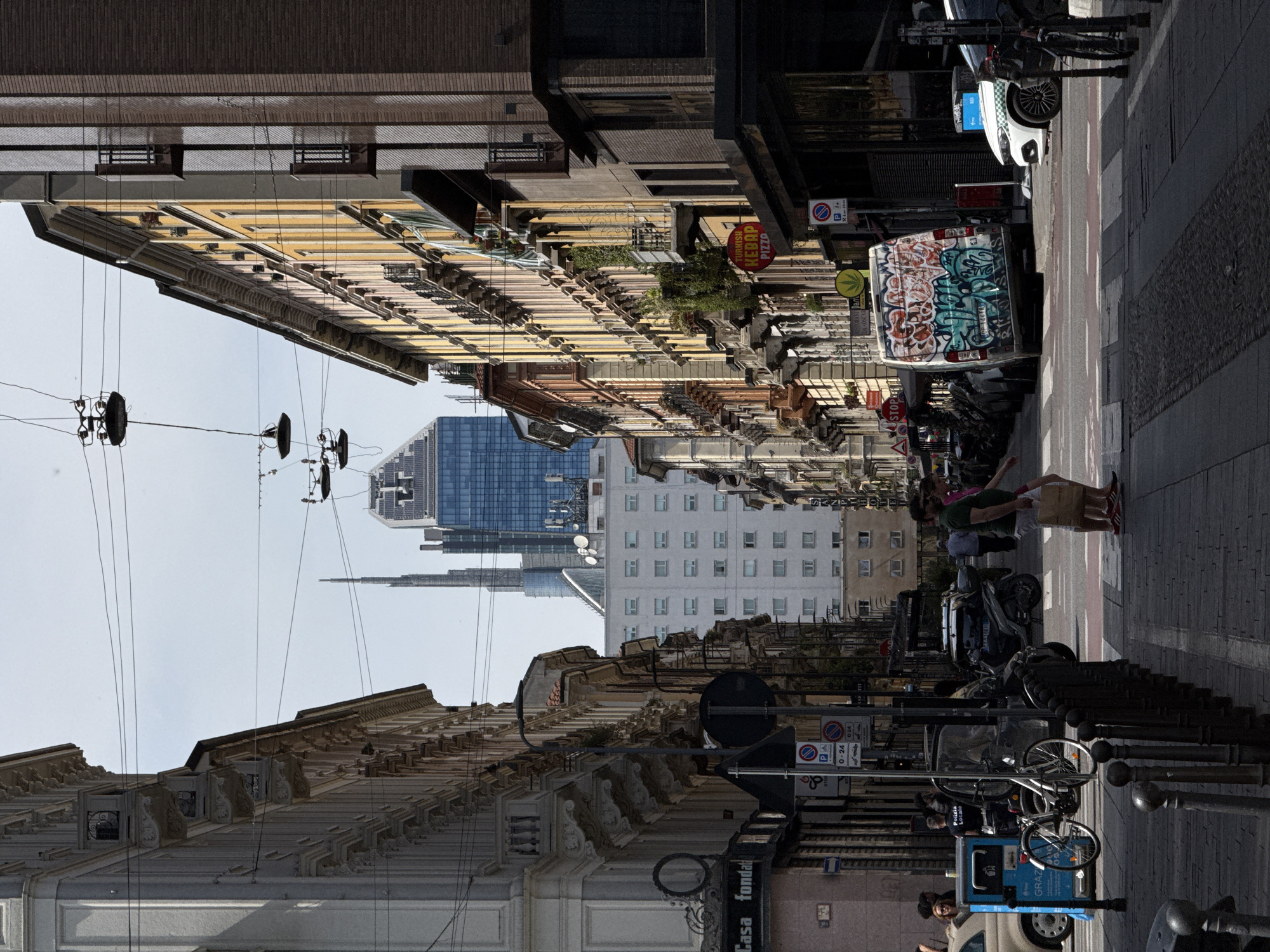 The image depicts a street scene in Milan, Italy. The perspective is taken from within the street, looking towards a cluster of skyscrapers in the distance. Buildings line both sides of the street, featuring a mix of architectural styles, including ornate facades and more modern designs. Overhead, a network of power lines is visible against the sky. On the right side of the street, a van is parked and covered in graffiti. Also on the right, pedestrians are walking along the sidewalk, and bicycles are parked. Street signs are visible along the sides of the street. The scene is brightly lit, suggesting a sunny day.