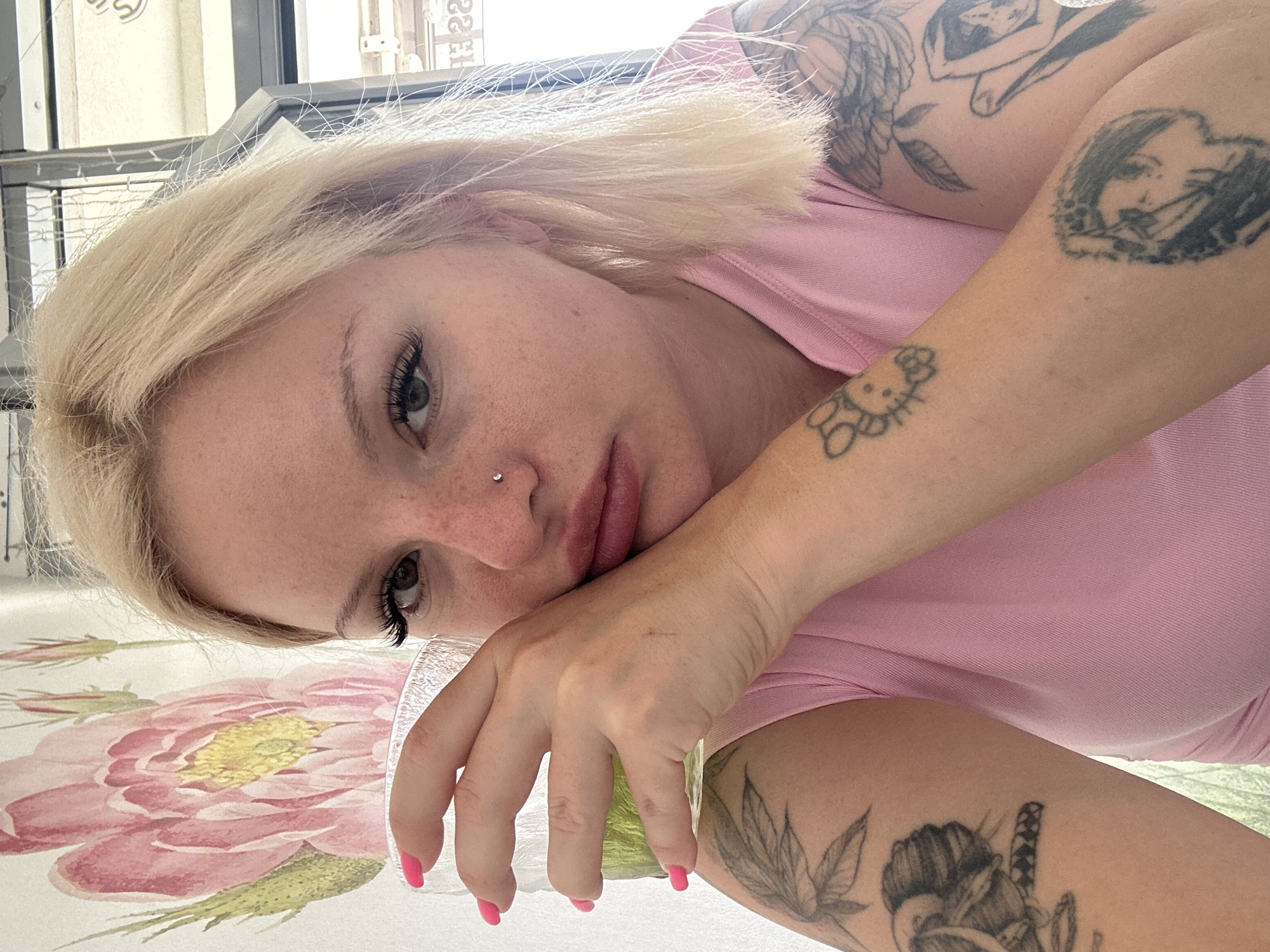 A young woman with blonde hair and a nose piercing holds a glass of green liquid in her right hand. She is wearing a pink t-shirt and has tattoos on both arms, including a Hello Kitty design on her left arm and a stylized portrait on her right arm. She is leaning on her hand, with her elbow on a table, and her face is close to the glass. The background shows a floral print wall and a window with a blurred view of a street. This appears to be an outdoor cafe in Budva, Montenegro, possibly near the Old Town.
