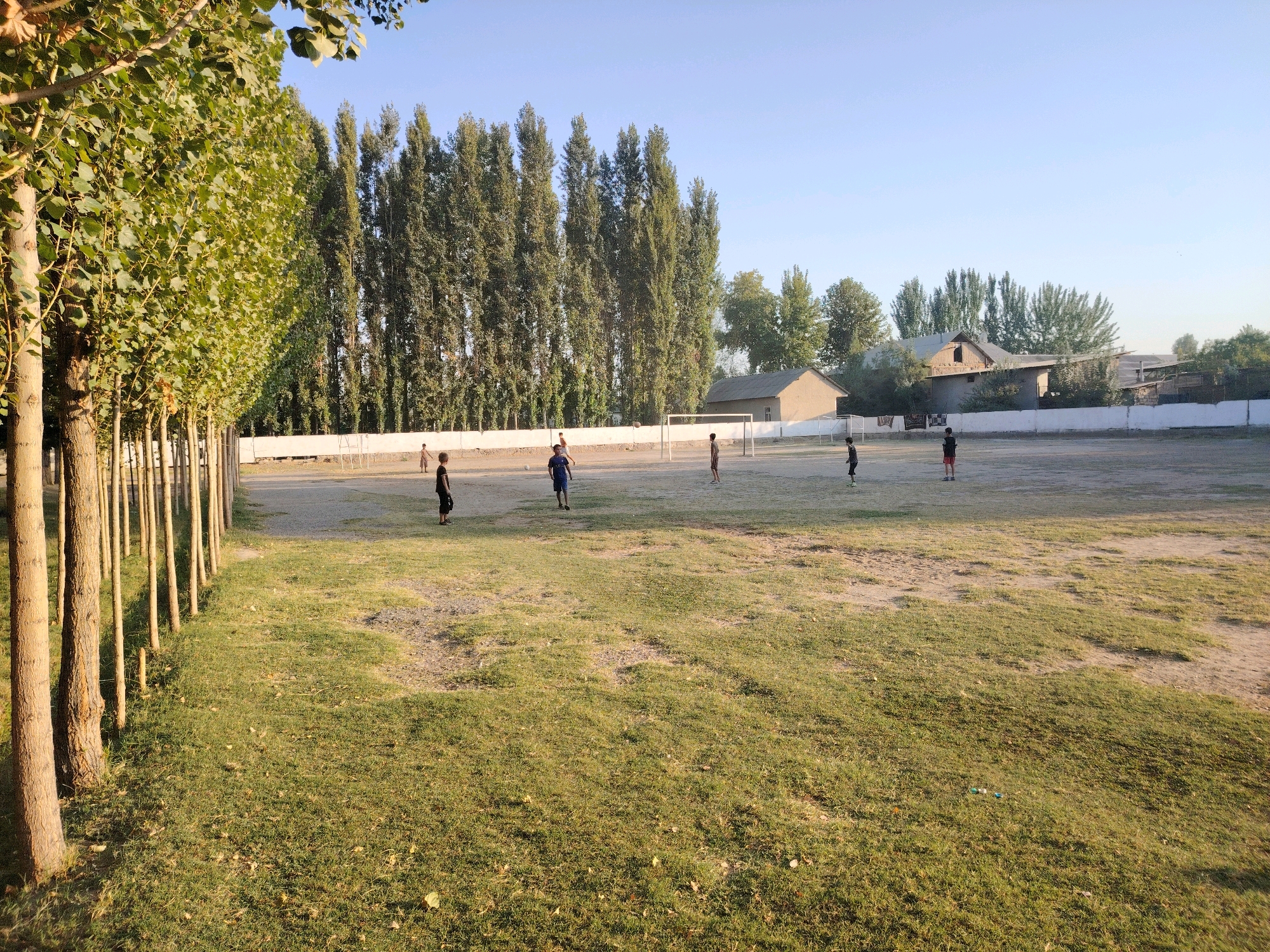 The image depicts an outdoor scene in the city of Paxtaobod, Uzbekistan, likely on a sports field or park. In the foreground, a grassy area with patches of dirt is visible, with a line of tall trees on the left casting shadows. In the mid-ground, several children are scattered across a dusty field, some appearing to be playing soccer. Two soccer goals are set up on opposite ends of the field. In the background, buildings and more trees are visible under a clear blue sky. The lighting suggests it is either early morning or late afternoon.