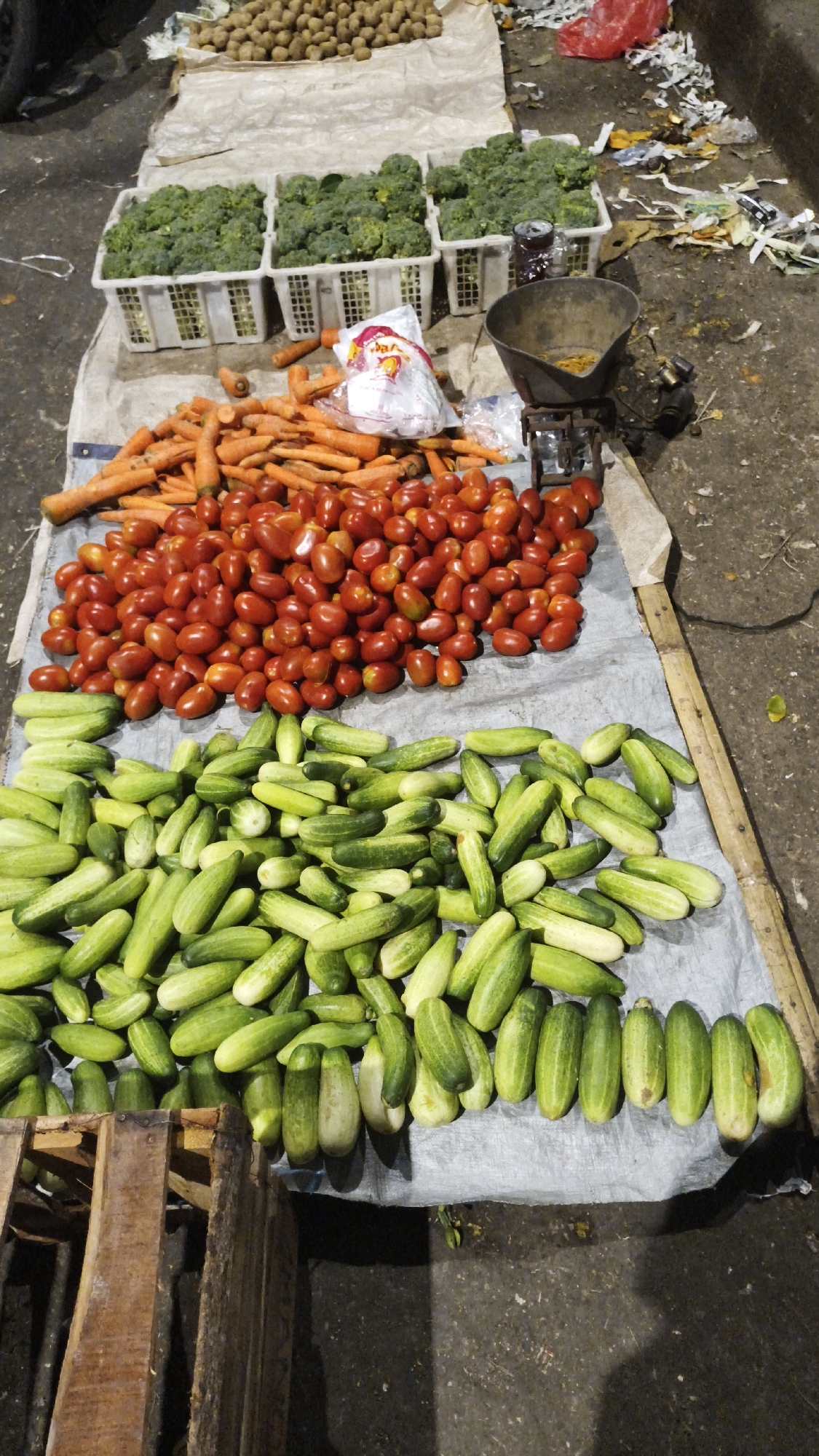 At a street market in South Jakarta, Indonesia, a selection of fresh produce is displayed on a grey tarp. In the foreground, a large pile of green cucumbers is neatly arranged. Behind the cucumbers, a mound of red cherry tomatoes is visible. Further back, a pile of orange carrots is spread out. Three white baskets containing broccoli are placed in the middle ground. To the right, an old metal scale with a dark bowl sits on the tarp, next to some trash. The ground is a rough, dark surface, possibly asphalt or concrete, and there are some discarded materials and wrappers scattered around the scene. A wooden crate is partially visible in the lower left corner. The lighting suggests it may be nighttime or a dimly lit area.