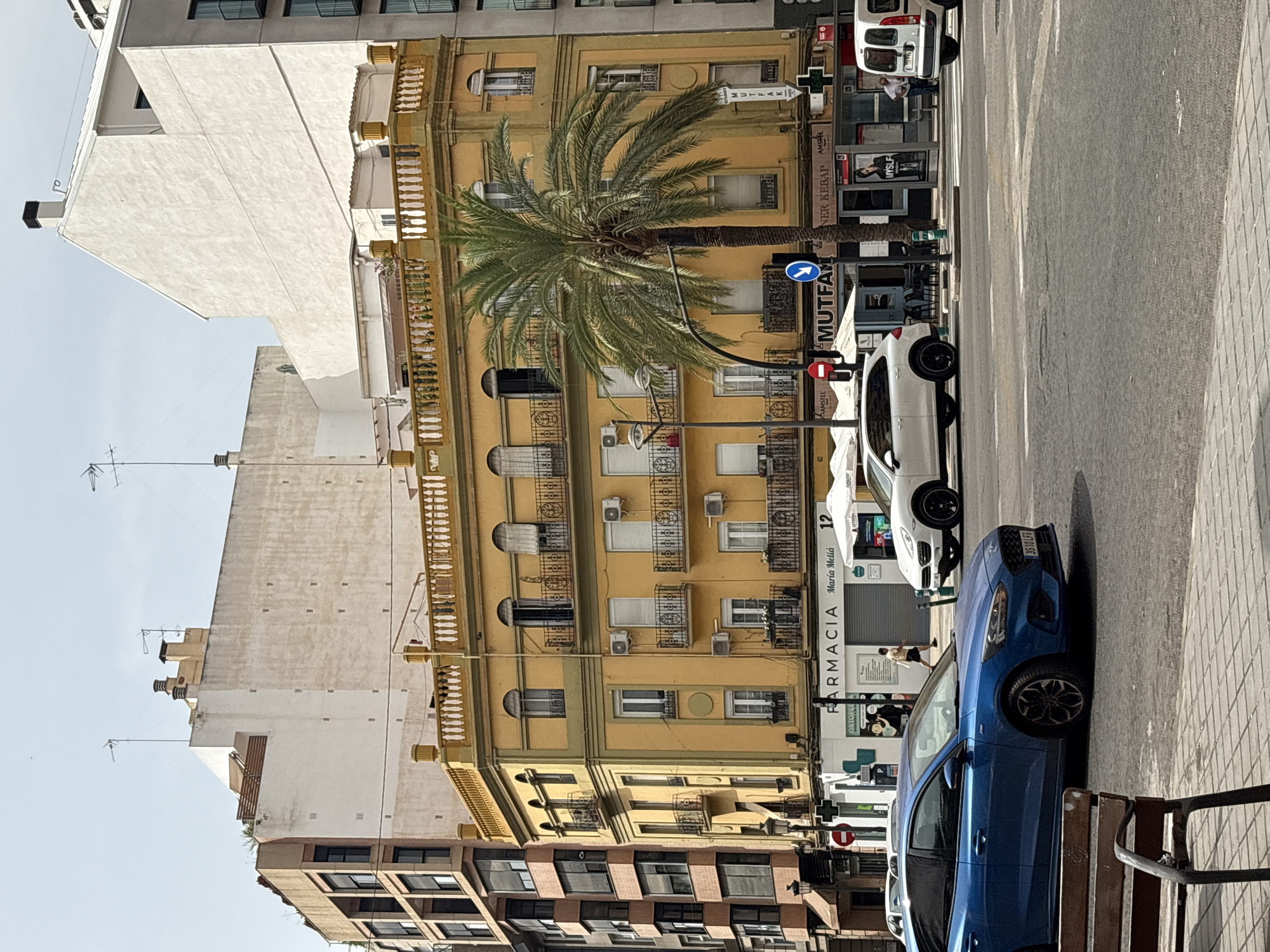 The image depicts a street scene in Valencia, Spain, on a sunny day. In the foreground, a blue Ford Focus is parked on the left side of the street. Behind it, a white SUV is seen in motion. Across the street, a yellow building with balconies and ornate railings stands prominently. A large palm tree grows in front of this building. Below the yellow building, a "Farmacia Maria Melia" is visible, along with other businesses. The background features several multi-story buildings with varying architectural styles. Pedestrians are present on the sidewalk, and traffic signs and streetlights are also visible. The overall impression is of a busy urban environment in Valencia.