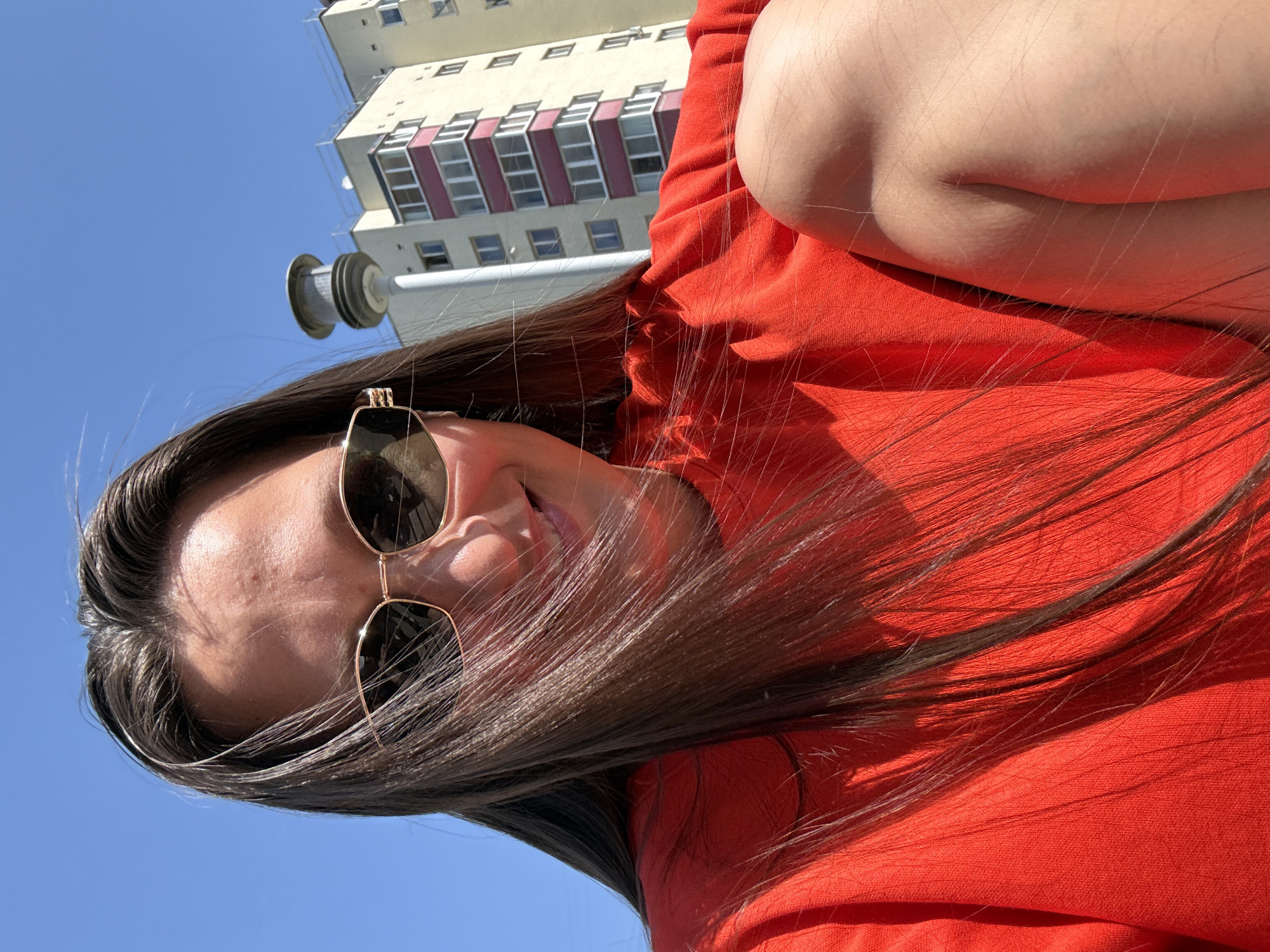 A low-angle, portrait-style photograph features a woman wearing sunglasses and a bright red shirt. A light-colored, multi-story building with a repeating pattern of windows (some with red frames) is partially visible in the background, along with a cylindrical object attached to a pole. The sky is bright blue. The location is likely in Chaikovsky, Russia, based on the request.