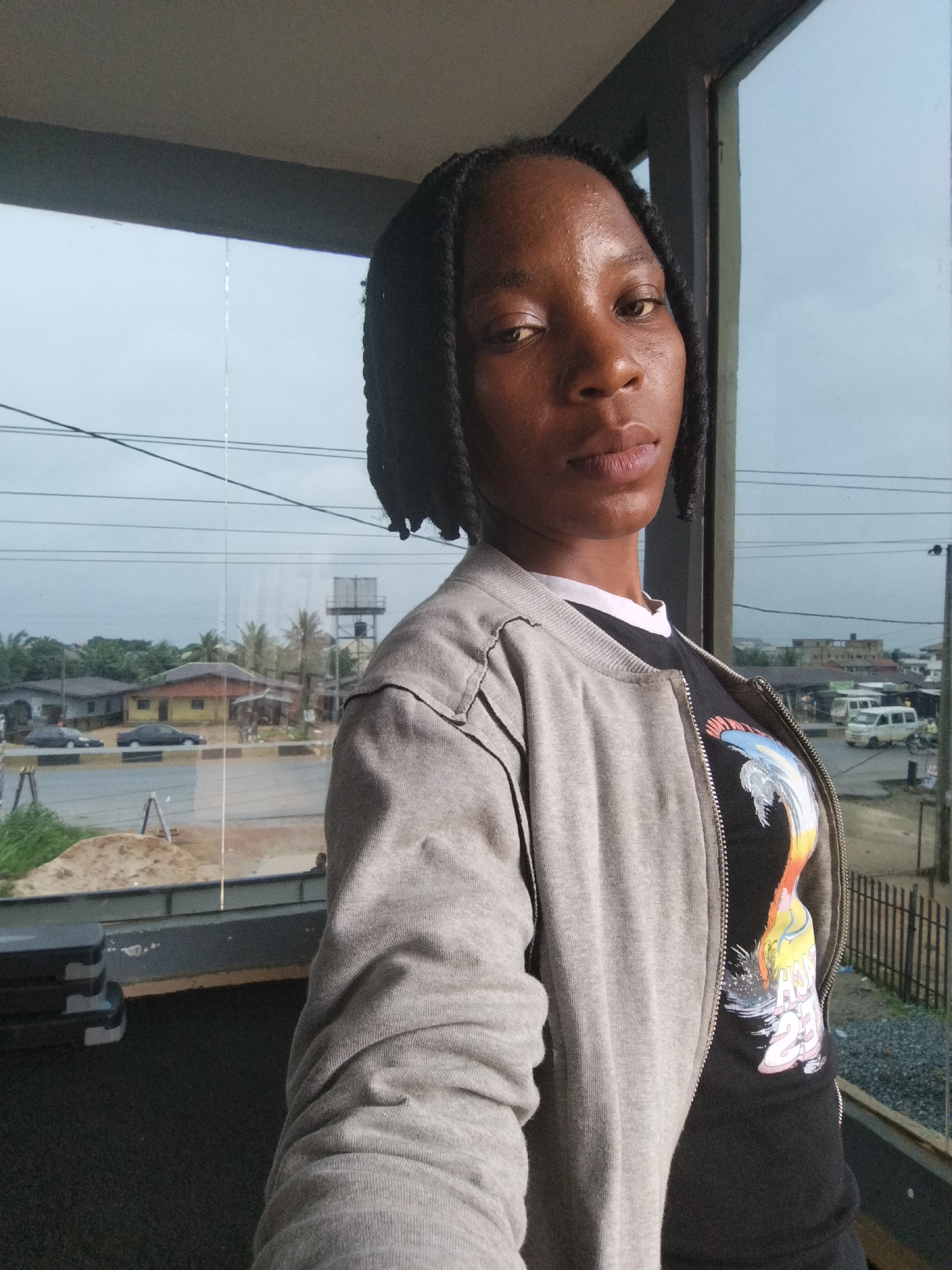 A young Black woman with dark skin and dark braided hair is looking directly at the camera. She is wearing a gray zippered jacket over a black t-shirt with a colorful graphic design on it. The woman is standing indoors, in front of a large window that looks out onto a street scene in Uselu, Nigeria. The exterior shows houses, palm trees, cars, and a water tower. The lighting suggests it is daytime, but the sky is overcast.