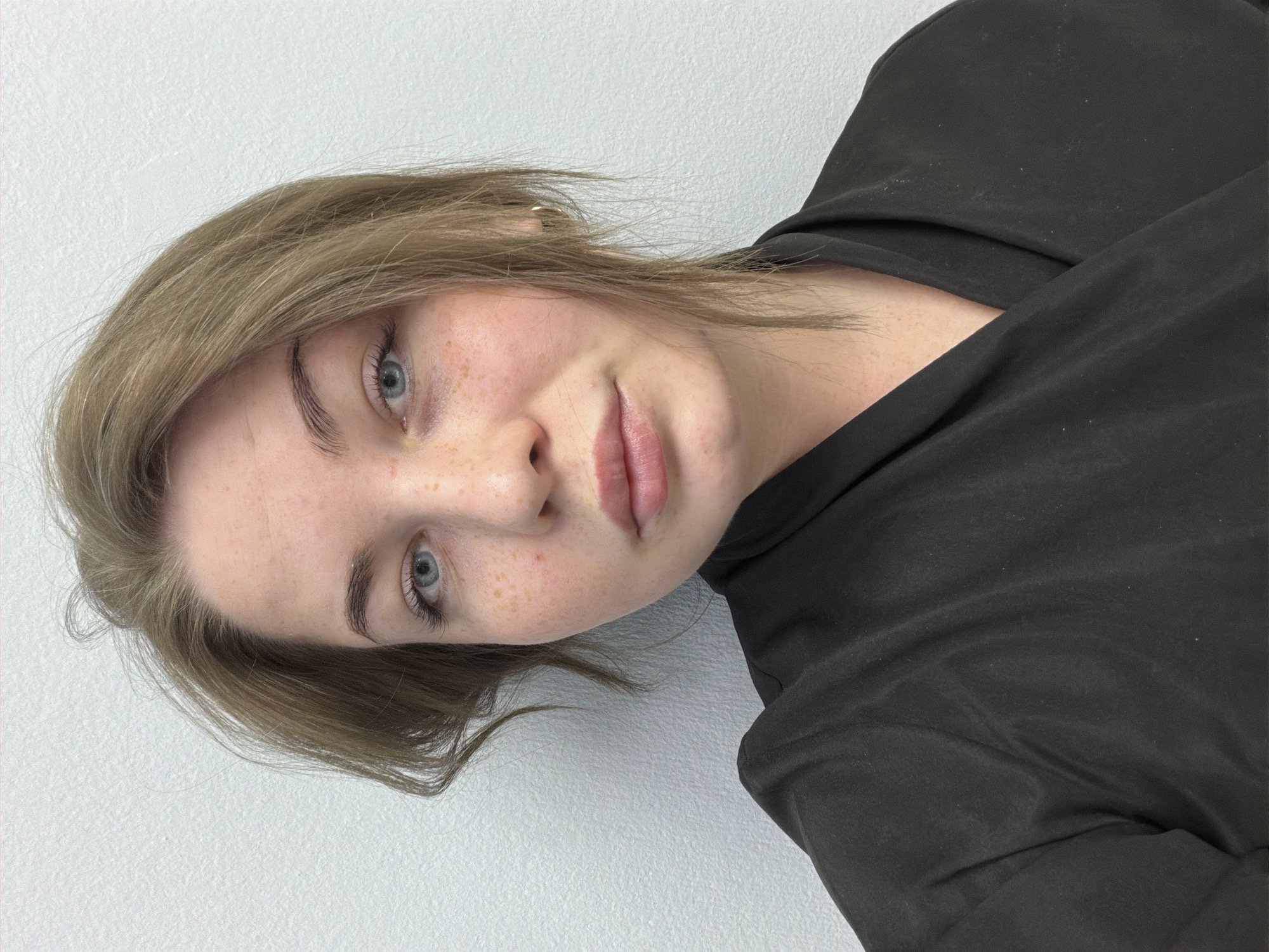 The image shows a close-up, waist-up portrait of a young woman with light brown hair and blue eyes, facing the camera. She has fair skin with freckles and is wearing a black wrap-style top. The background is a plain, textured white wall. The lighting is even and appears to be artificial. There is no indication of the city of Chaikovsky, Russia.