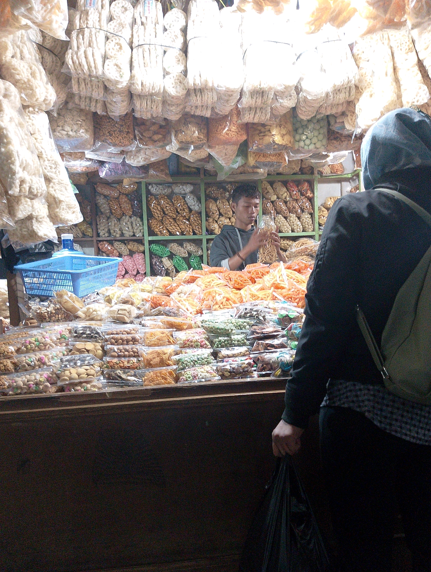 The image captures a bustling stall in Lembang, Indonesia, likely a traditional market given the variety of packaged snacks and treats. The stall is filled with an abundance of food items, primarily snacks, displayed in clear plastic packaging. These packages are arranged on shelves and laid out on a counter.
In the background, a young man is attending to the stall. He is wearing a grey hooded sweatshirt and is holding up a package of what appear to be dried noodle-like snacks. The shelves behind him are stocked with more packaged goods, including what look like nuts, crackers, and brightly colored candies.
In the foreground, a person wearing a dark hooded jacket and a light-colored checkered shirt is standing with their back to the camera, holding a black plastic bag. Their right hand is visible, gripping the bag. The person appears to be either browsing the stall or preparing to make a purchase.
The stall itself is densely packed with merchandise, creating a vibrant and visually stimulating scene. Various types of snacks are visible, including puffed snacks, nuts, dried fruits, and what appear to be small, round candies in different colors. A blue plastic basket is also visible on the counter. The overall impression is one of abundance and a lively market atmosphere in Lembang.