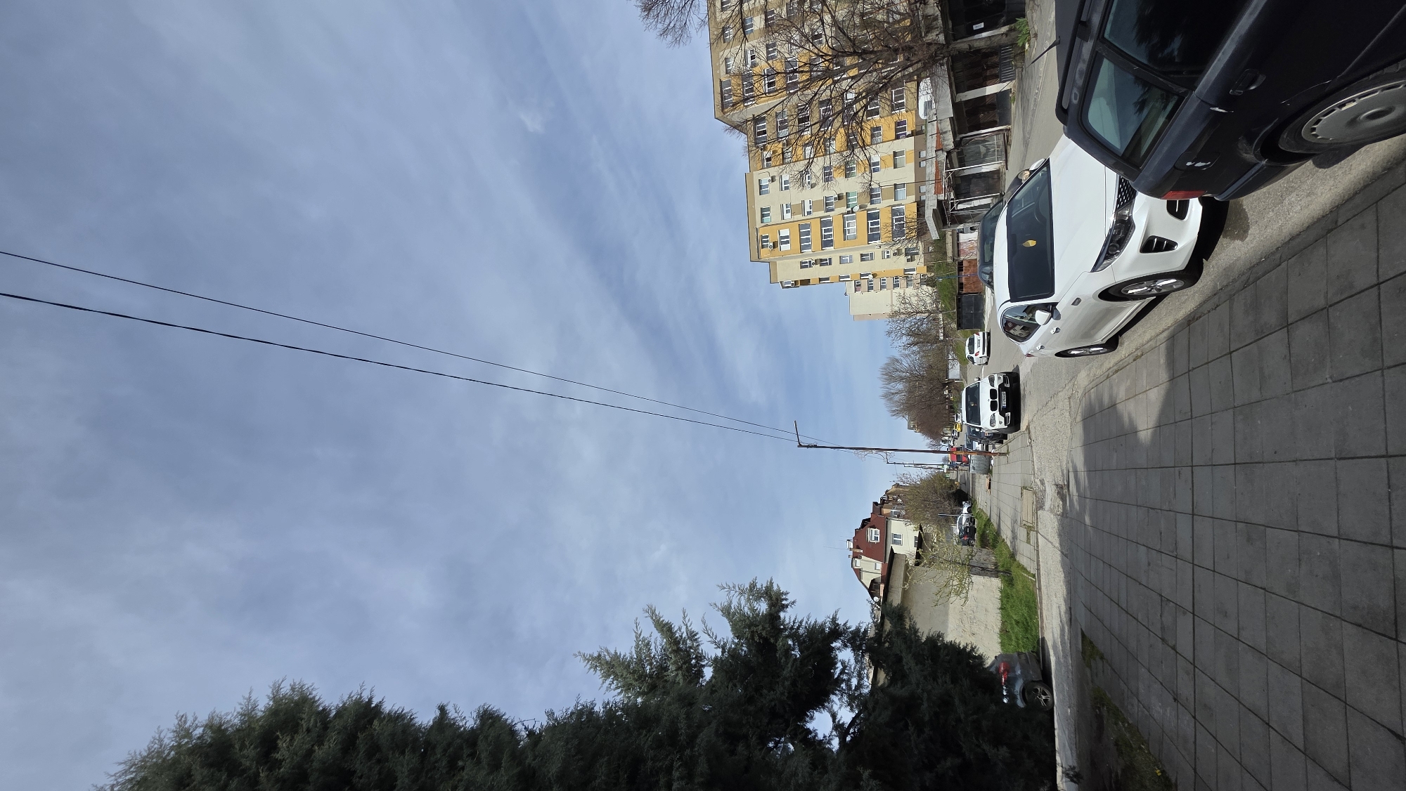 The image shows a street scene in Blagoevgrad, Bulgaria. The photograph appears to be taken from a low angle, looking up the street.
**Environment:** The sky is partly cloudy. The street is lined with buildings of varying heights and architectural styles, including a large apartment block. The street has a tiled pavement with cars parked along it. Trees are visible along the side of the street.
**Objects:** The main objects are buildings, cars (including white and black vehicles), a street light post, overhead power lines, and trees. The apartments are multiple stories.
**Subjects:** The image includes a view of everyday urban life. There are no people discernible.
