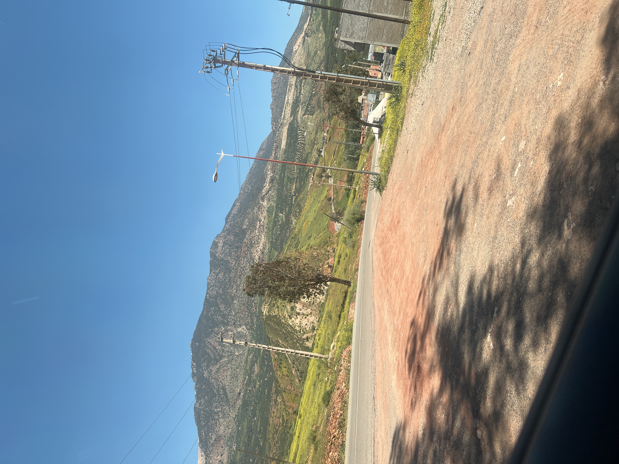 The image depicts a rural roadside scene in Ouaouizert, Morocco. A paved road runs along the side of a mountain, with a dirt shoulder. Power lines and utility poles stretch along the roadside and through the landscape. A single tree stands along the slope. In the background, the mountain is visible with varied greenery. The sky is clear and blue. Tree shadows are cast on the dirt shoulder of the road. A portion of a vehicle interior is visible in the foreground.