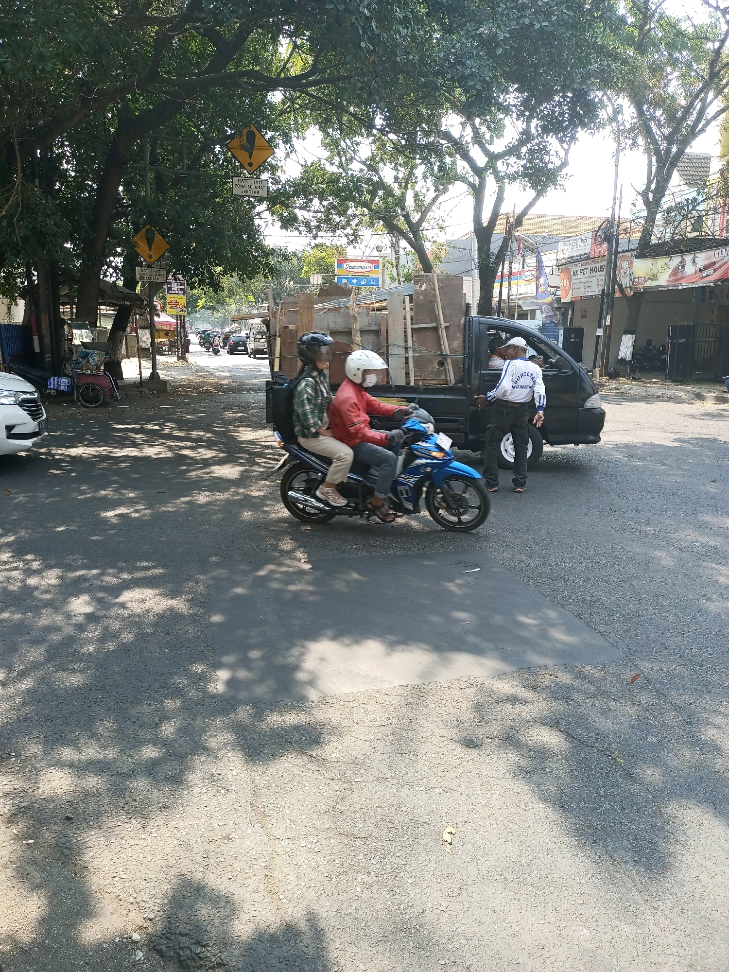 Two individuals wearing helmets are riding a blue motorcycle on a paved road. The person in the front is wearing a red jacket, and the person in the back is wearing a green and white checkered shirt. Behind them, a black truck is parked, with a person in a white shirt and dark pants standing next to it, facing the truck. The scene appears to be in a city in Malang, Indonesia, indicated by the street signs visible in the background. The environment includes trees casting shadows on the road, buildings with signage, and other vehicles in the distance. The lighting suggests it is daytime, likely sunny.