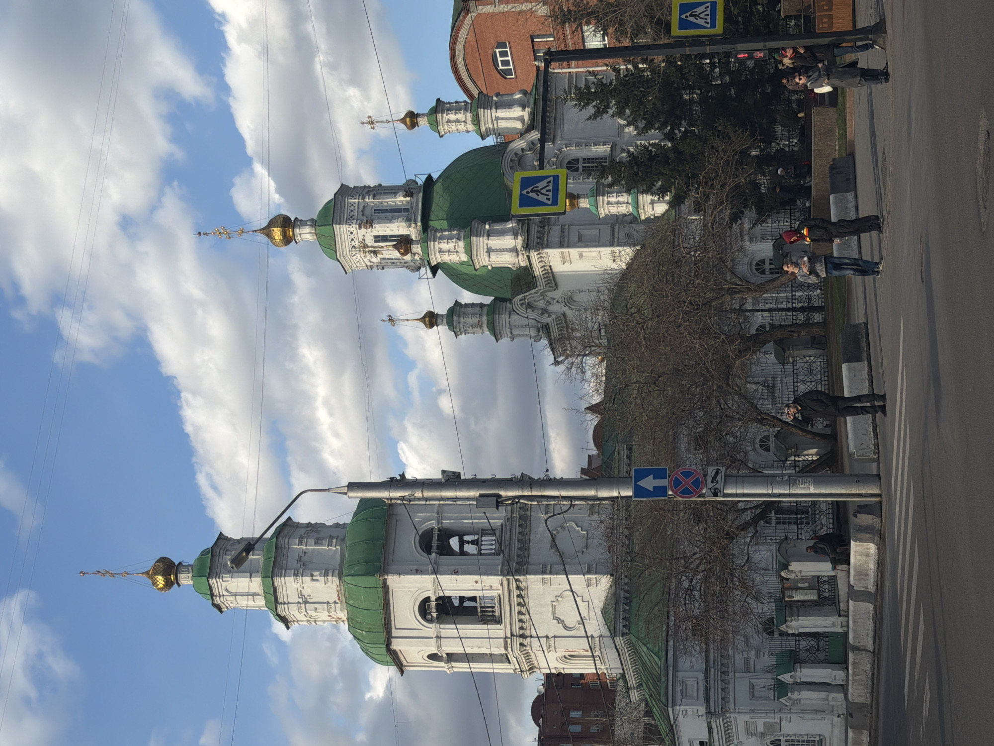 The image shows the Annunciation Cathedral in Krasnoyarsk, Russia. The cathedral's distinctive white walls, green domes, and golden crosses are prominently featured against a partly cloudy sky. Power lines are visible in the sky. Along the street adjacent to the cathedral, pedestrians are present. Street signs are also visible, including a pedestrian crossing sign and a no parking sign. The street appears to be asphalt. Buildings of varying heights and architectural styles are visible in the background, indicating an urban setting. Some bare branches of trees are also visible along the street.