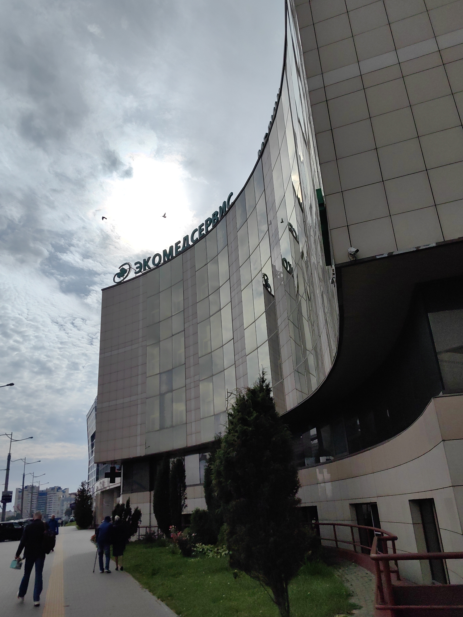 This is a low-angle shot of a modern, curved building with a facade of glass and light-colored panels, situated on a city street. The building has a prominent green logo and text that reads "ЭКОМЕДСЕРВИС" (Ecomedservice) at the top. The sky is overcast with patches of bright sunlight breaking through the clouds, and two birds are visible in the sky.
In the foreground, a sidewalk runs alongside the building, with a few people walking. On the left, a man in a dark jacket and blue jeans walks away from the camera, carrying a light-colored bag. Further down the sidewalk, a couple walks towards the camera; the man is using a walking stick. A few evergreen trees and some rose bushes are planted on a grassy area next to the sidewalk.
In the background, streetlights and other buildings are visible, indicating an urban environment. The overall scene suggests a typical day in the city. Based on the building's signage, this location is likely the Ecomedservice clinic in Minsk, Belarus.