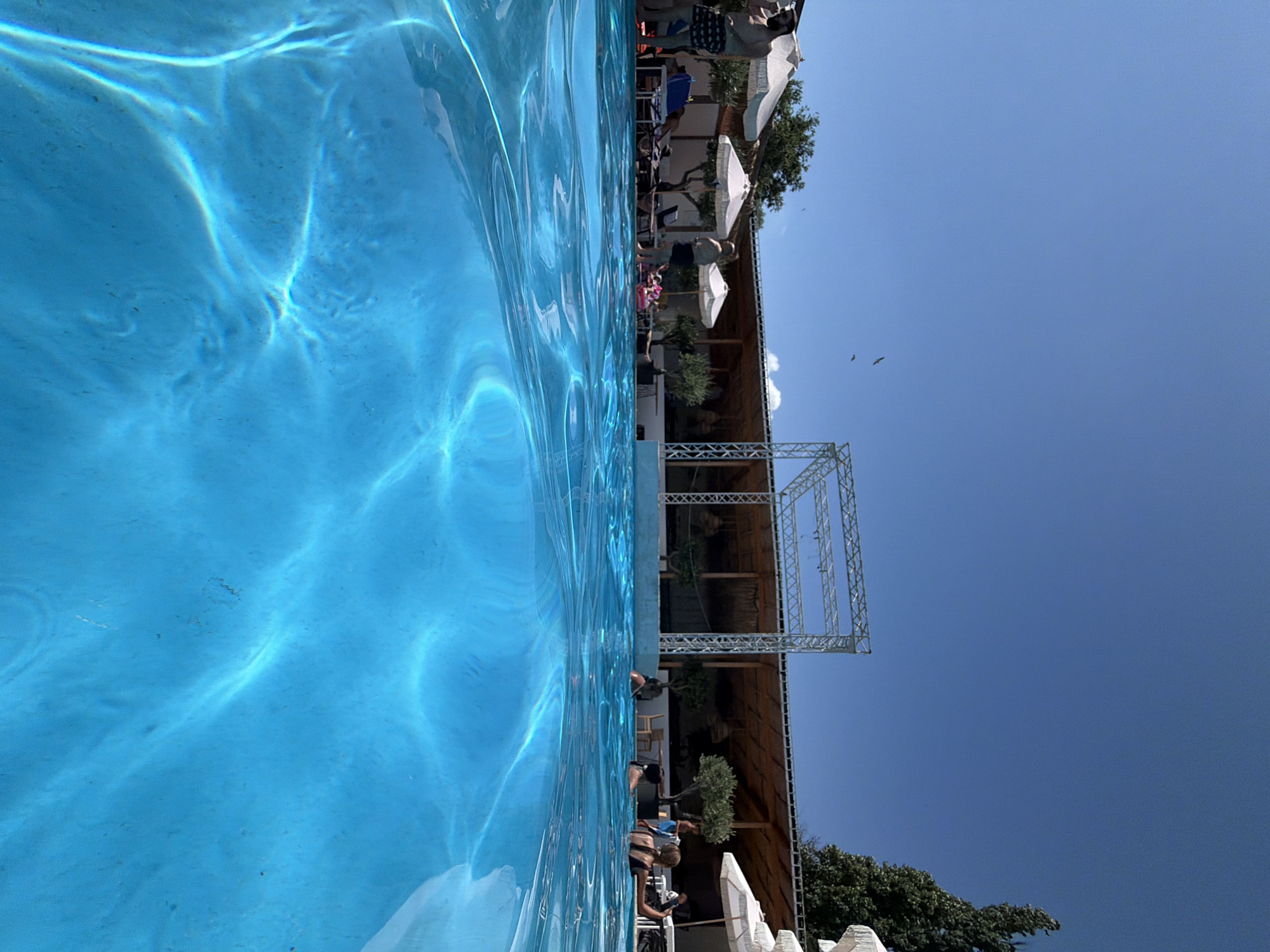 An underwater and above-water shot captures a bright, sunny day at a pool in Burgas, Bulgaria. The foreground is dominated by the clear, turquoise water of the swimming pool, with sunlight creating dancing patterns on the pool floor. In the background, people are visible relaxing and swimming. Several white umbrellas provide shade for lounge chairs. Behind the pool area, there is a covered structure with wooden beams and thatched roofing, suggesting a bar or restaurant area. A silver metal frame, possibly a stage or lighting rig, is set up in front of the covered structure. Trees and greenery are scattered throughout the scene. Two birds are flying in the clear blue sky. The overall impression is one of a vibrant and popular outdoor leisure spot.