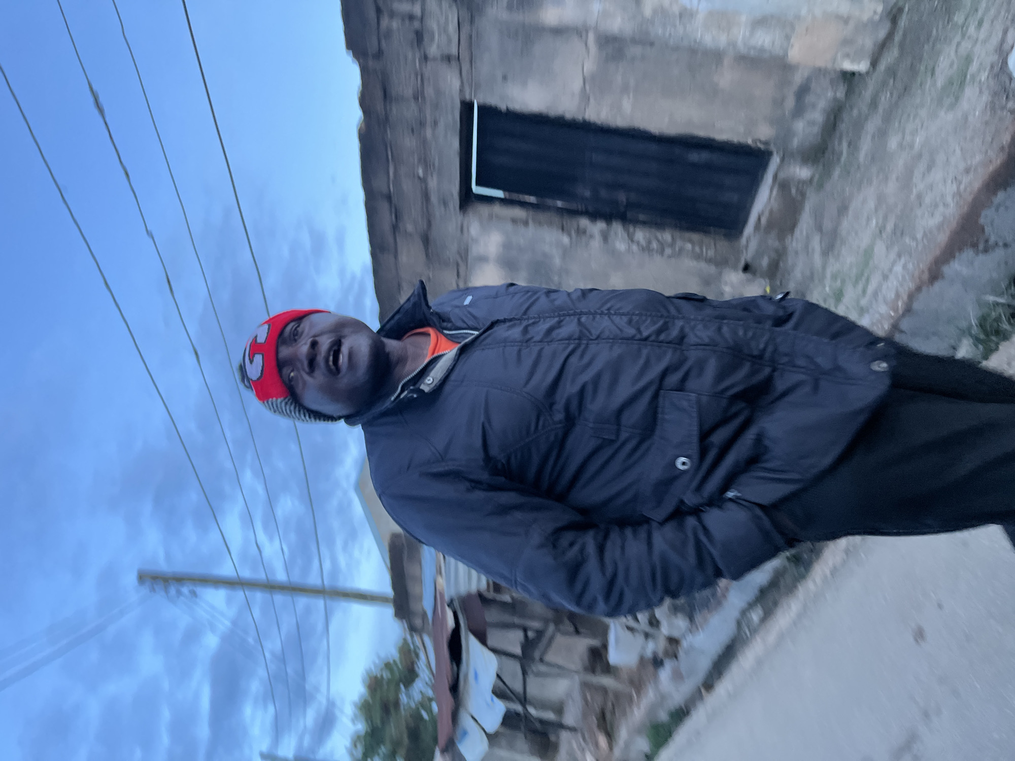 An adult male with dark skin stands outdoors in what appears to be dawn or dusk lighting, characterized by a muted blue-grey sky. He is positioned slightly to the viewer's right of the center, facing upwards and to his left, with his mouth slightly open. He wears a red and grey knit beanie with a white "G" logo, a dark blue or black quilted puffer jacket over an orange undershirt, and dark trousers. His hands are tucked into his jacket pockets.
The ground beneath him is an unpaved, light brown and grey dirt path, with a partial concrete curb visible on the left side.
In the midground, to the viewer's left of the man, are rudimentary structures featuring corrugated metal roofs, some secured by objects, and parts of a light-colored building with green foliage from trees or bushes visible further behind. A wooden utility pole with multiple sagging overhead wires runs diagonally from the mid-left upwards.
To the viewer's right of the man, also in the midground, stands a weathered, grey concrete or masonry building with a dark, rectangular doorway opening on its right side.
The upper portion of the image is dominated by a blue-grey, cloudy sky. The scene depicts a residential or semi-rural area in the city of Unknown, Unknown.