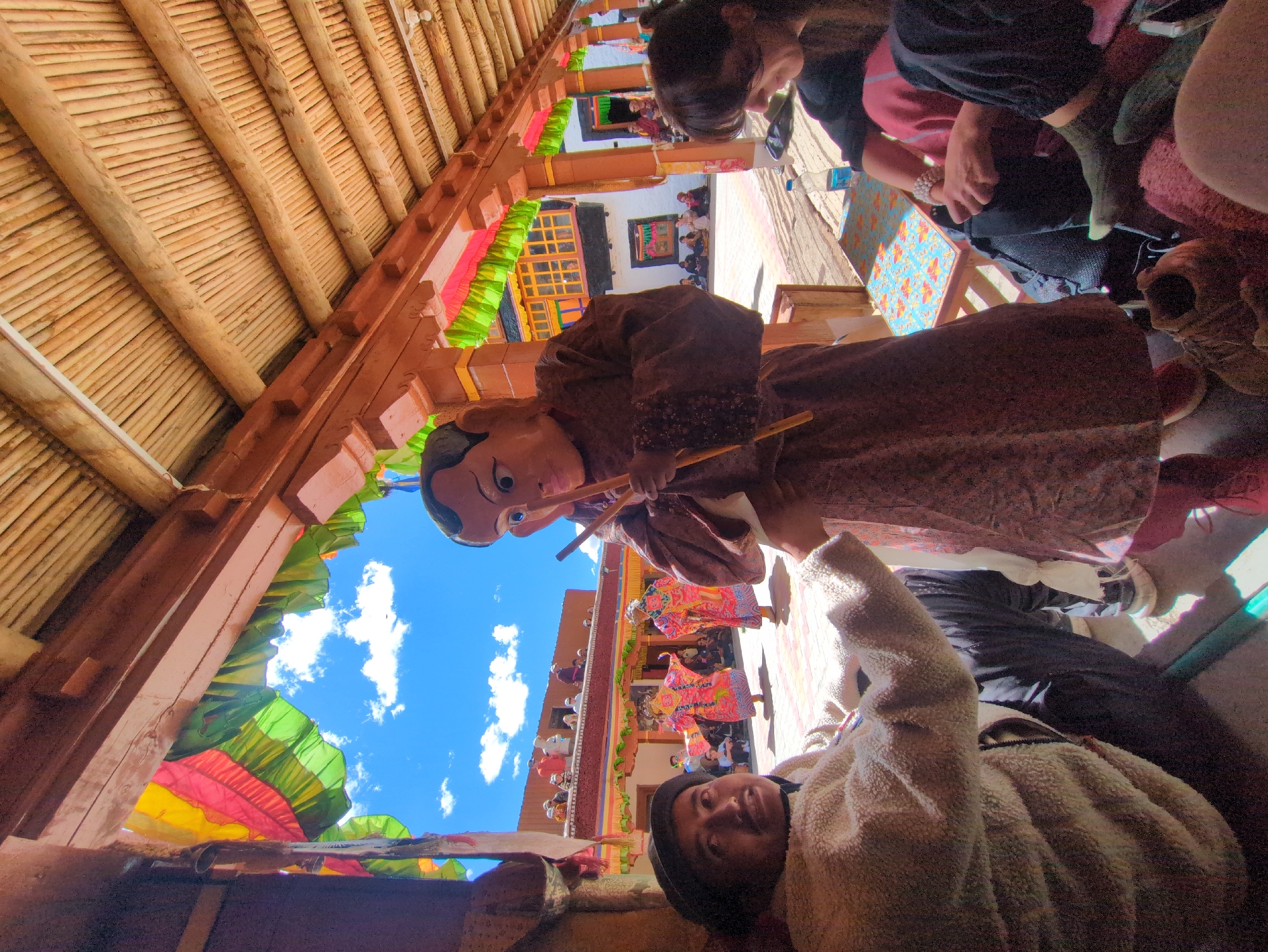 In the city of Pibiting, India, a lively outdoor gathering is underway, possibly at a monastery, as indicated by the architectural style and the presence of people in traditional attire. The scene is framed by a wooden roof structure supported by pillars, adorned with colorful fabric banners in red, yellow, and green. Below, a person in a beige, fuzzy jacket sits facing forward, looking up with a slight smile. In front of them, a figure wearing a large, mask-like headpiece with wide eyes and a pointed nose stands holding two wooden sticks. This figure is clad in a brown robe. In the background, two individuals in bright, ornate pink and yellow costumes are performing a dance, their movements captured mid-motion. A crowd of spectators sits or stands around the courtyard, observing the festivities. The sky above is a brilliant blue, dotted with white clouds, suggesting a clear, sunny day. The ground appears to be paved.