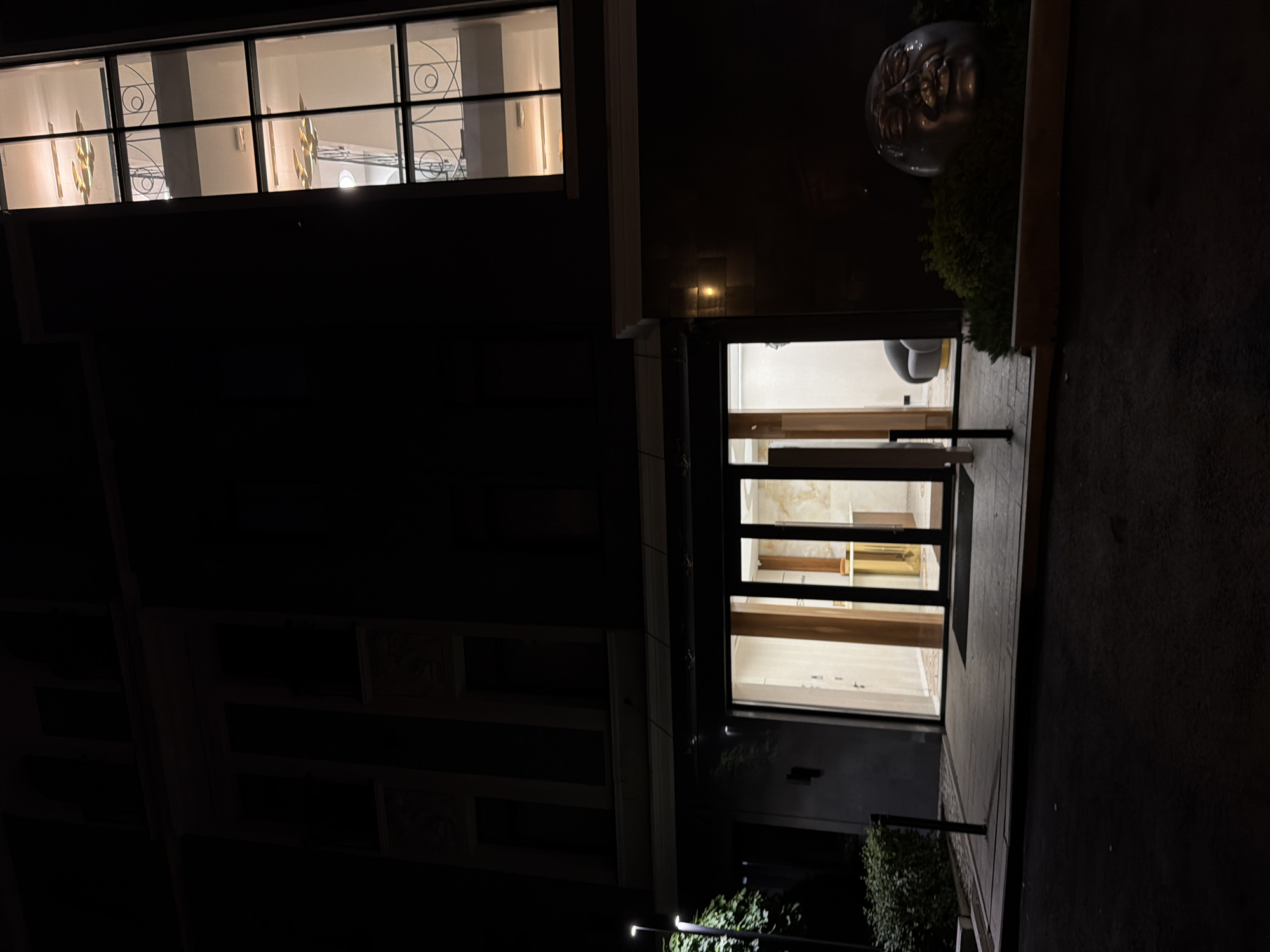 A nighttime exterior shot captures a dark, modern building with lit windows and an unusual sculpture. The building's facade is predominantly dark, with a tall, narrow window on the right side illuminating interior spaces with distinct floor levels, featuring ornamental railings and modern light fixtures. Below this, a wide glass entranceway glows with interior light, revealing a reception area with wood paneling, a counter, and glass doors. To the right of the entrance, nestled in a planter with green foliage, is a large, metallic sculpture resembling a human head with an exaggerated, somewhat distressed facial expression. In the foreground, a paved walkway leads to the entrance, with a streetlamp visible on the left side of the image, casting light on some surrounding greenery and the pavement. The overall scene is one of understated, contemporary architecture contrasted with an intriguing artistic element.