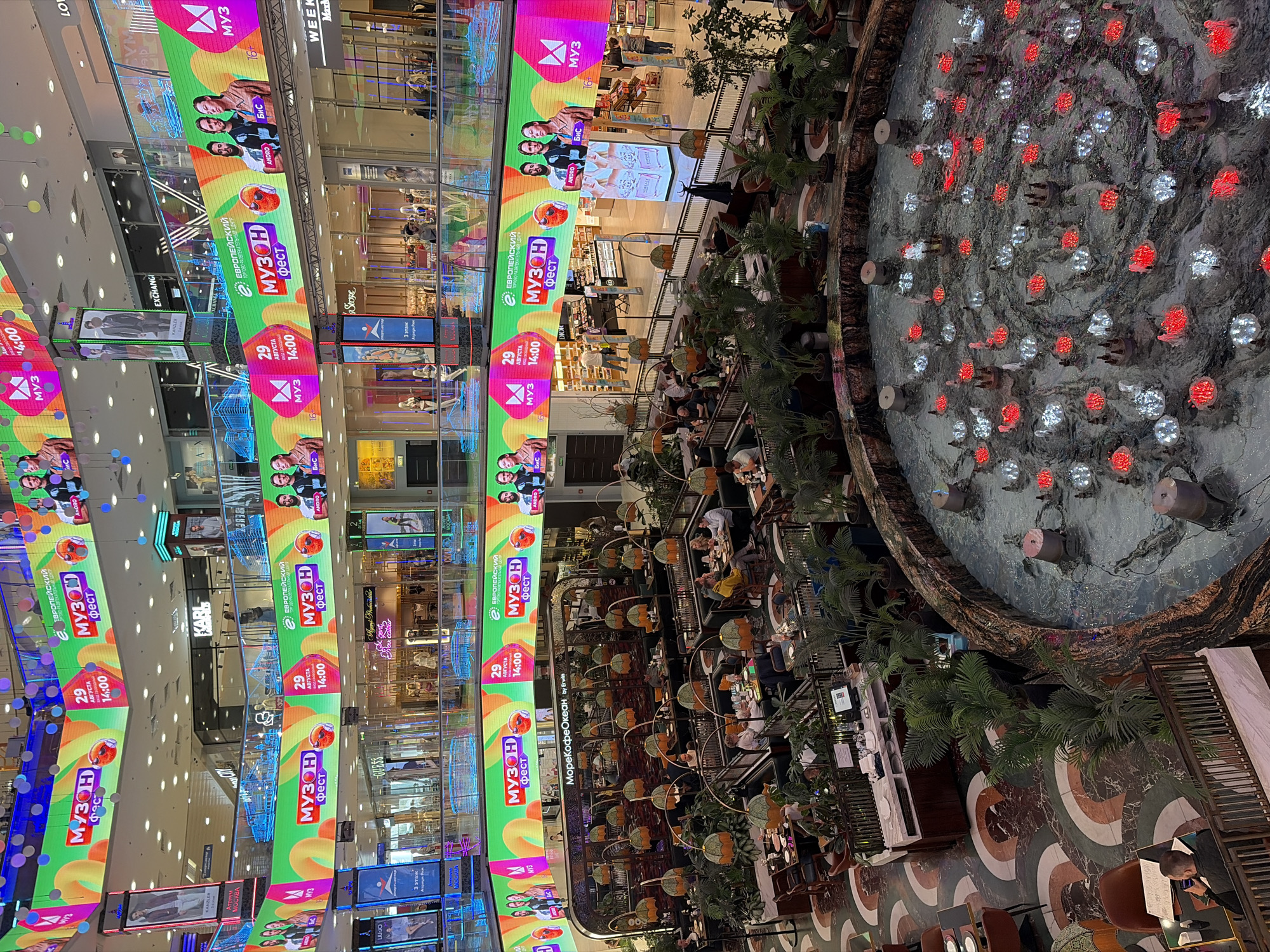 The image shows a multi-level interior of a large shopping mall, likely in Moscow, Russia, identified by Russian text on the digital billboards. The upper levels feature expansive, brightly lit digital screens displaying advertisements for a "MUZON FEST" on August 29th at 14:00. The screens show images of people, musical notes, and various graphical elements. The mall's architecture is modern, with multiple floors connected by escalators and walkways, and shops lining the lower levels.
In the foreground, a circular fountain is visible, with water jets and colorful lights (red and white). Surrounding the fountain is a large restaurant area with numerous tables and chairs. Diners are seated at several tables, engaged in conversations or eating. Lush green plants are strategically placed throughout the restaurant and surrounding areas, creating a biophilic design element. The floor of the mall features a decorative marble pattern. People are seen walking on different levels, shopping, or dining. The overall atmosphere appears to be a busy, commercial environment with a focus on entertainment and leisure.