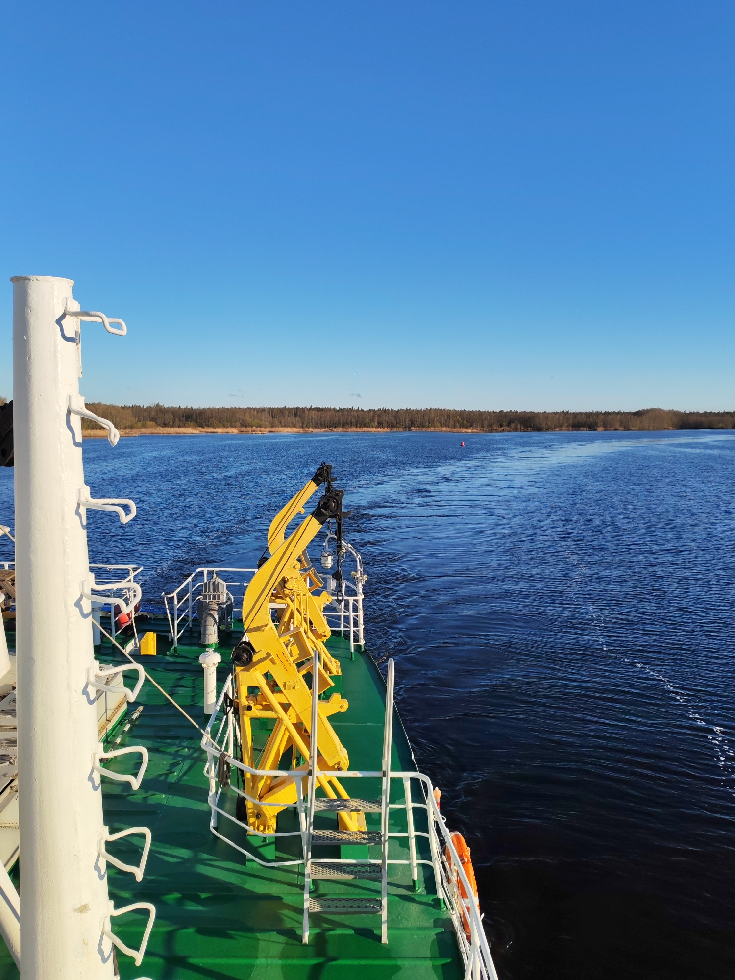 View from a ship on a waterway in Zubovo, Russia. The ship deck is green with white railings and includes yellow equipment. In the distance is a treeline under a clear blue sky. A red buoy is visible in the water. The waterway reflects the blue sky.