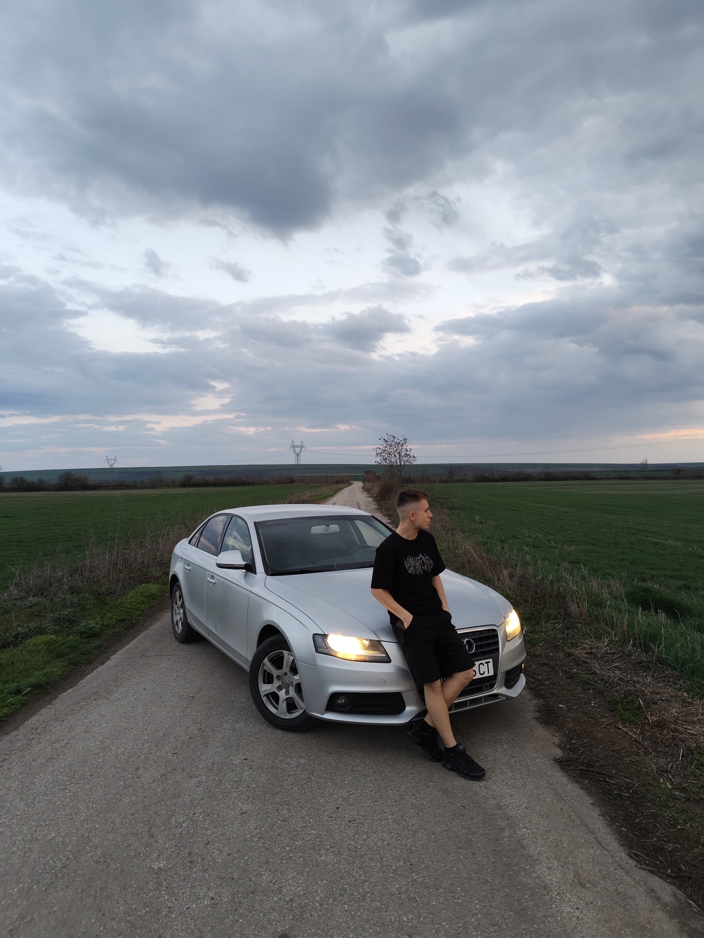 The image portrays a rural road in Gorni Tsibar, Bulgaria, under an overcast sky with predominantly gray clouds. A silver Audi A4 sedan is parked diagonally on a narrow, asphalt road, facing toward the viewer's right. Its headlights are illuminated. A young adult male is leaning against the front hood of the vehicle, facing right and looking away from the camera. He wears a black short-sleeved t-shirt with a white graphic, black shorts, and black athletic shoes. His right hand is in his pocket, and his left arm rests on the car's hood. The car's front license plate is partially visible, displaying "5 CT". Green agricultural fields border both sides of the road. In the mid-distance, a bare tree stands near a slight curve in the road. Further in the background, two high-voltage electricity pylons are visible against the horizon.