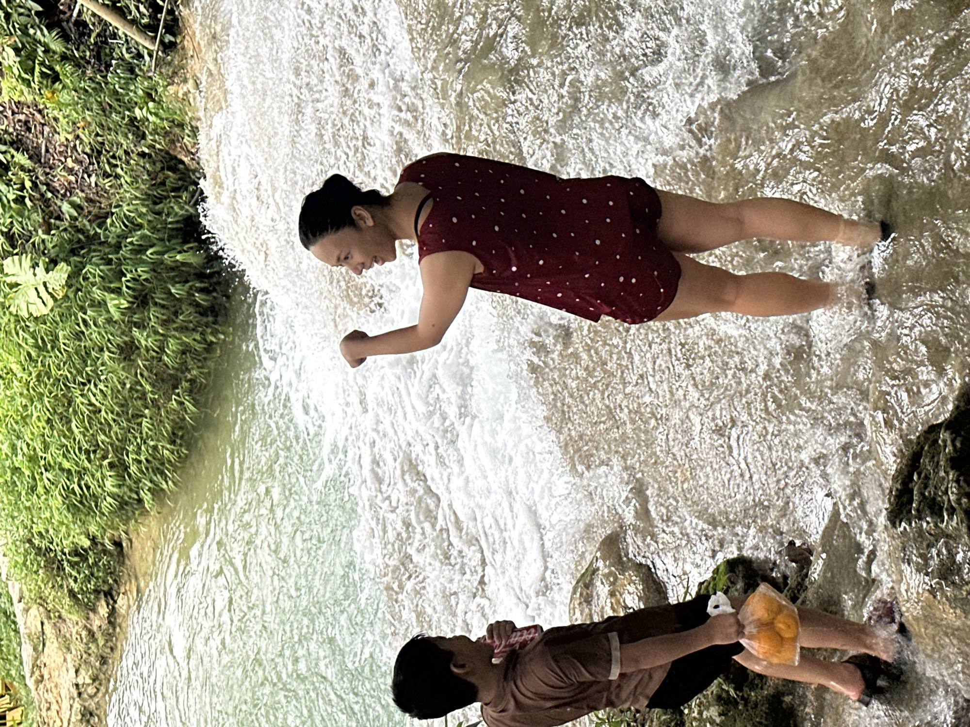 Two individuals are standing in shallow, rushing water at the base of a waterfall, likely in Cổ Lũng Commune, Vietnam, surrounded by lush green foliage and rocky terrain. A young boy, on the left, wears a brown t-shirt and black shorts, holding a clear plastic bag filled with several orange fruits. To his right, a woman in a dark red, polka-dotted sleeveless top and matching shorts faces away from the camera, her arm raised and her mouth open as if speaking or laughing. The water is turbulent and foamy, particularly around the woman. The environment suggests a natural, outdoor setting, possibly a tourist attraction given the presence of people and the preserved state of the foliage.