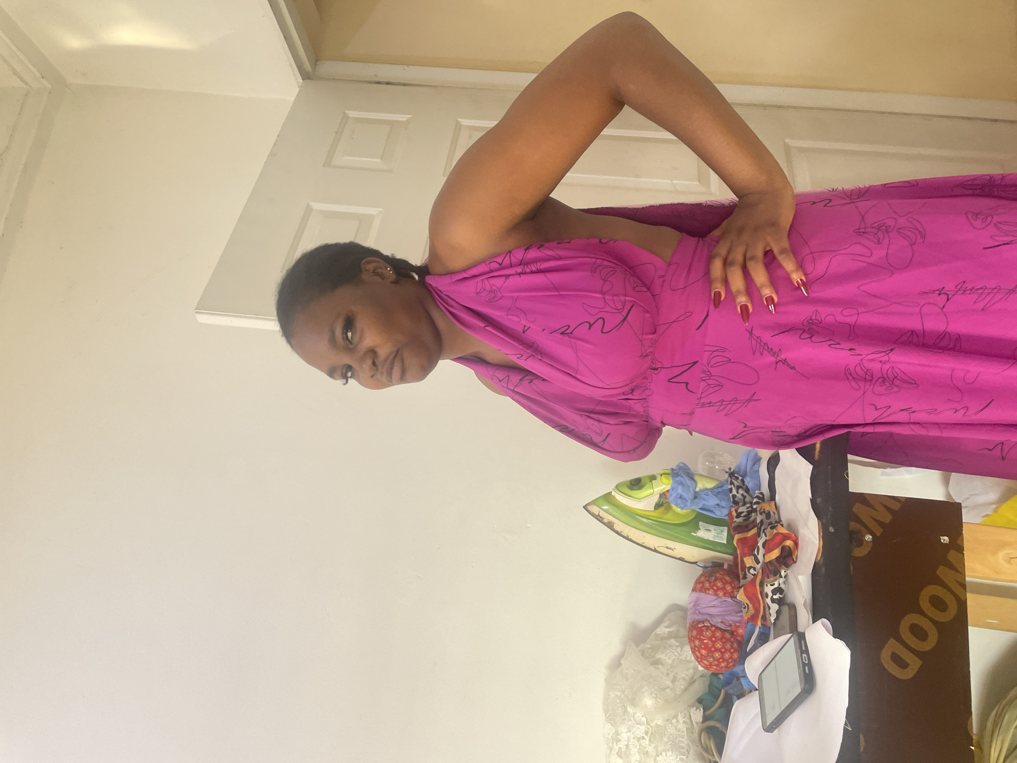 The image features an adult Black woman posing indoors in Dushepe, Nigeria. She is depicted from the waist up, looking directly at the camera with a neutral expression. Her dark hair is pulled back, and she wears small earrings. She is dressed in a vibrant fuchsia-colored halter-neck dress with abstract black line patterns. Her right arm is bent, and her hand, adorned with long red fingernails, rests on her hip.
The background consists of plain light-colored walls and a white door with two rectangular panels. To her right, a surface holds an assortment of objects. A green, vintage-style iron with some blue fabric draped over it is visible, alongside various piles of colorful textiles and clothing. A black smartphone with a blank screen rests on a piece of white fabric. Below these items, a dark brown wooden board displays the word "WOOD" in yellow capital letters. The setting appears to be an indoor domestic space, possibly a bedroom or dressing area.