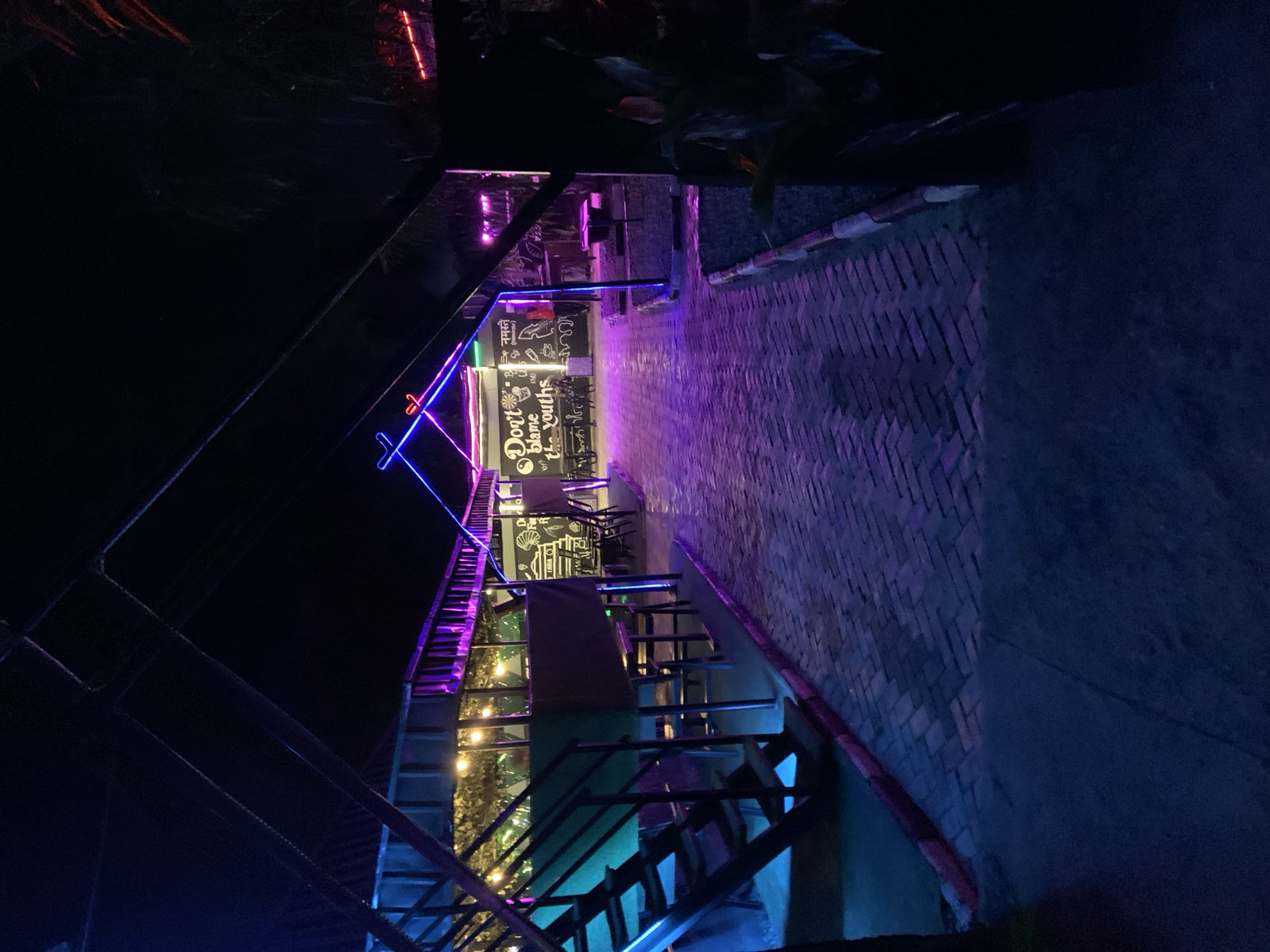 The image depicts an outdoor seating area of a restaurant or cafe at night, illuminated by colorful neon lights and string lights. The ground is paved with brick, and there are tables and chairs set up for customers. A staircase leading to an upper level is visible on the left side of the image. The overall ambiance is festive and inviting, with the neon lights casting vibrant hues of blue and purple across the scene. The establishment appears to be located in Gura Top, Nigeria, suggesting a vibrant nightlife scene in the city.