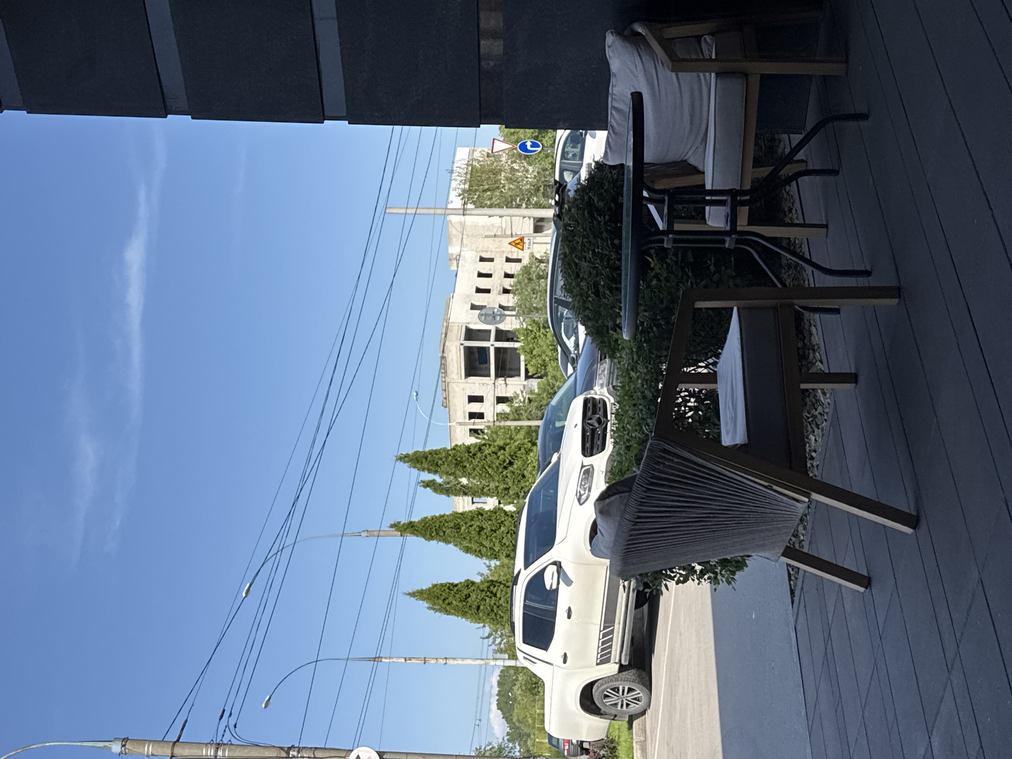 The image shows an outdoor seating area with furniture, including a table and chairs, positioned on a deck. Beyond the seating area, a white pickup truck is parked, and several cypress trees are visible. In the background, there is a partially constructed building and power lines crisscrossing the bright blue sky. The scene suggests a sunny day, possibly in the late morning or early afternoon, given the clear sky and bright lighting. The overall atmosphere is calm and relaxed, typical of an outdoor cafe or a residential patio. There is no visible text in the image.