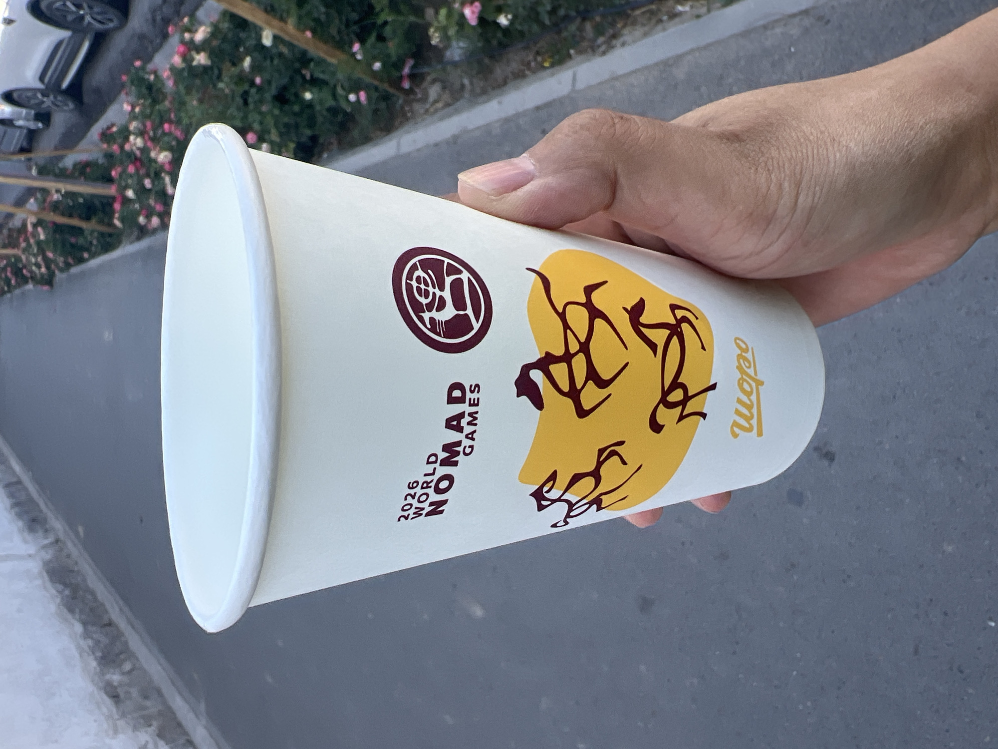 In Osh City, Kyrgyzstan, a person's hand holds a white paper cup with a maroon logo and text. The cup features the text "2026 WORLD NOMAD GAMES" in maroon, with stylized dark brown horse silhouettes on a yellow background below. A circular maroon emblem is also visible on the cup. The cup is held in front of a paved pathway lined with rose bushes and trees, with a white wall in the background. Partially visible vehicles are in the background on the right.