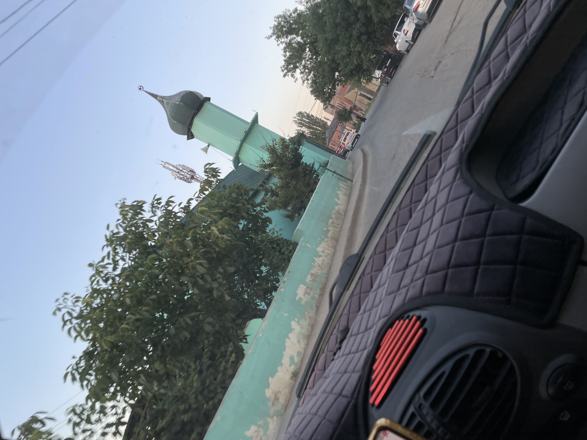 The image shows a street scene in Buynaksk, Russia, with a prominent turquoise-colored mosque with a green dome on the left side of the frame. A tall, slender minaret of the mosque extends into the clear blue sky. Trees with lush green foliage line the street, and the turquoise wall of the mosque, appearing somewhat weathered and peeling in places, runs along the left side. In the foreground, the interior of a vehicle is visible, including the dashboard with a quilted black cover and a red grille vent. Several cars are parked and driving on the asphalt road. The overall impression is a snapshot of urban life in Buynaksk, capturing architectural elements and the everyday flow of traffic.