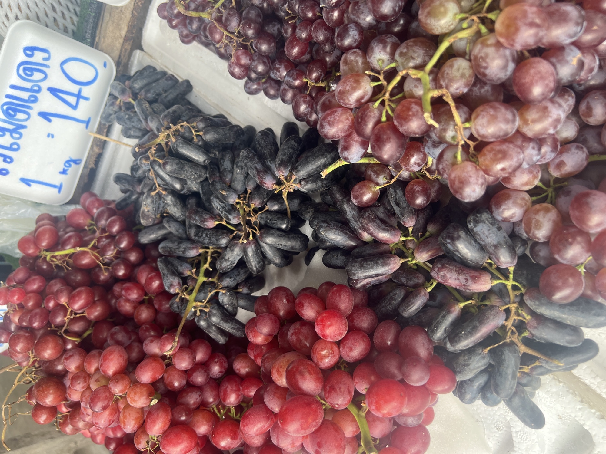 A close-up shot displays a selection of fresh grapes at a market stall, likely in the city of Ko Samui District, Thailand. In the foreground, three distinct varieties of grapes are arranged. To the left, a cluster of round, red grapes is visible. In the center, a bunch of elongated, dark purple grapes, often referred to as "moon drops," is prominently featured. To the right, another bunch of round, deep red or purple grapes is displayed. A white price tag with blue markings is partially visible in the upper right corner, indicating a price of "140" per kilogram for what appears to be the moon drop variety. The grapes are presented on a white styrofoam surface, suggesting a market or produce stand setting. The lighting is bright, highlighting the textures and colors of the fruit. No people or other consumers are present in the frame.