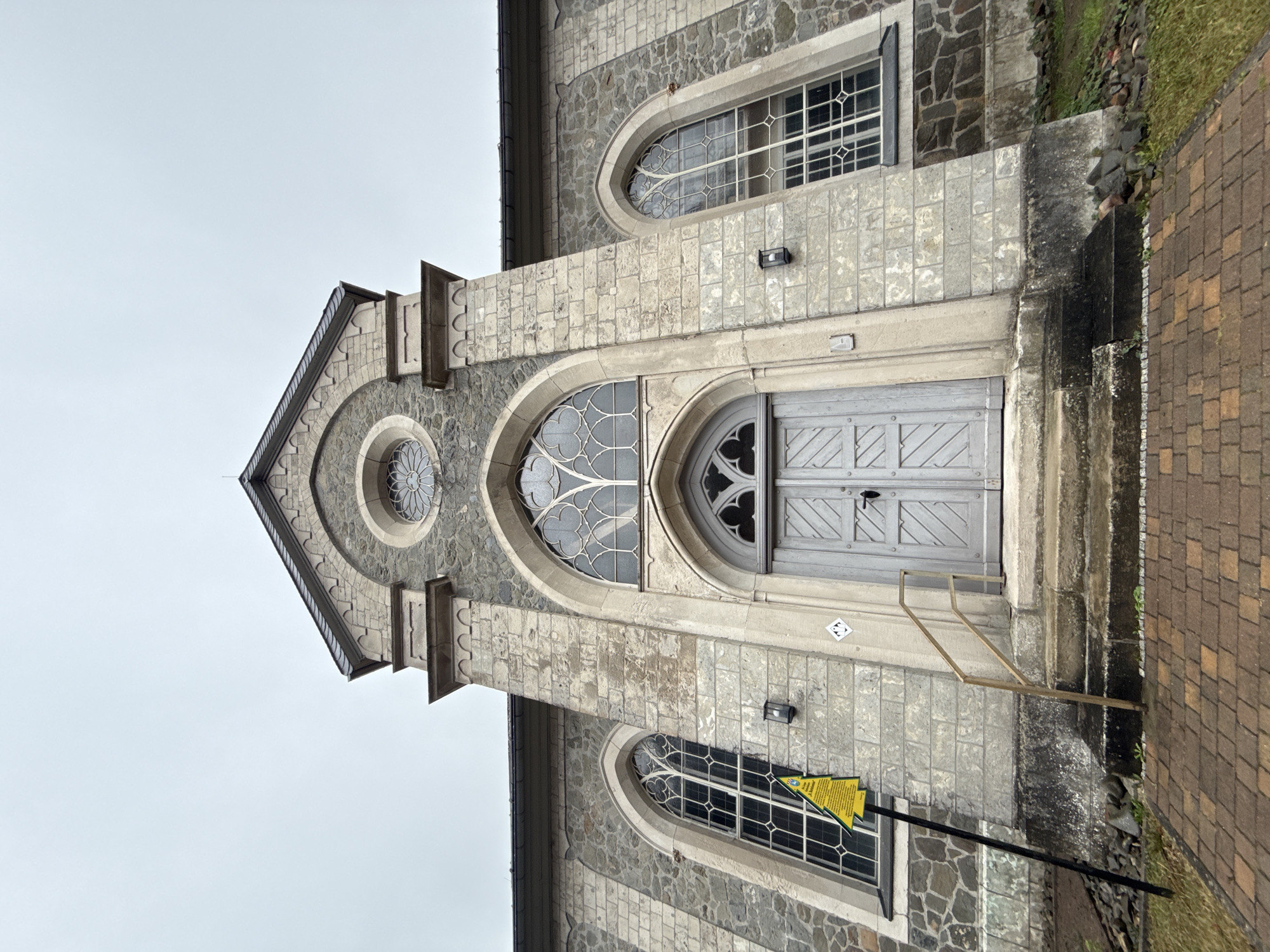 The image depicts the exterior of a stone church in Walkenried, Germany. The perspective is low-angle, looking up at the central entrance and facade. The church has a pointed arch doorway with double wooden doors painted light gray, and above it, a large arched window with intricate tracery. A circular rose window is situated above this. Flanking the central entrance are two tall, narrow arched windows, also with decorative tracery. The church is constructed of rough-hewn grey stone blocks with lighter stone quoins and architectural details. A dark gray roof is visible at the top of the structure. A yellow triangular sign with black text and an image of a person walking is mounted on a black pole to the left of the entrance. Two light fixtures are mounted on the facade near the large arched windows. Steps lead up to the entrance, and a metal handrail is present. The ground in front of the church is paved with reddish-brown brick. The sky is overcast and light gray.