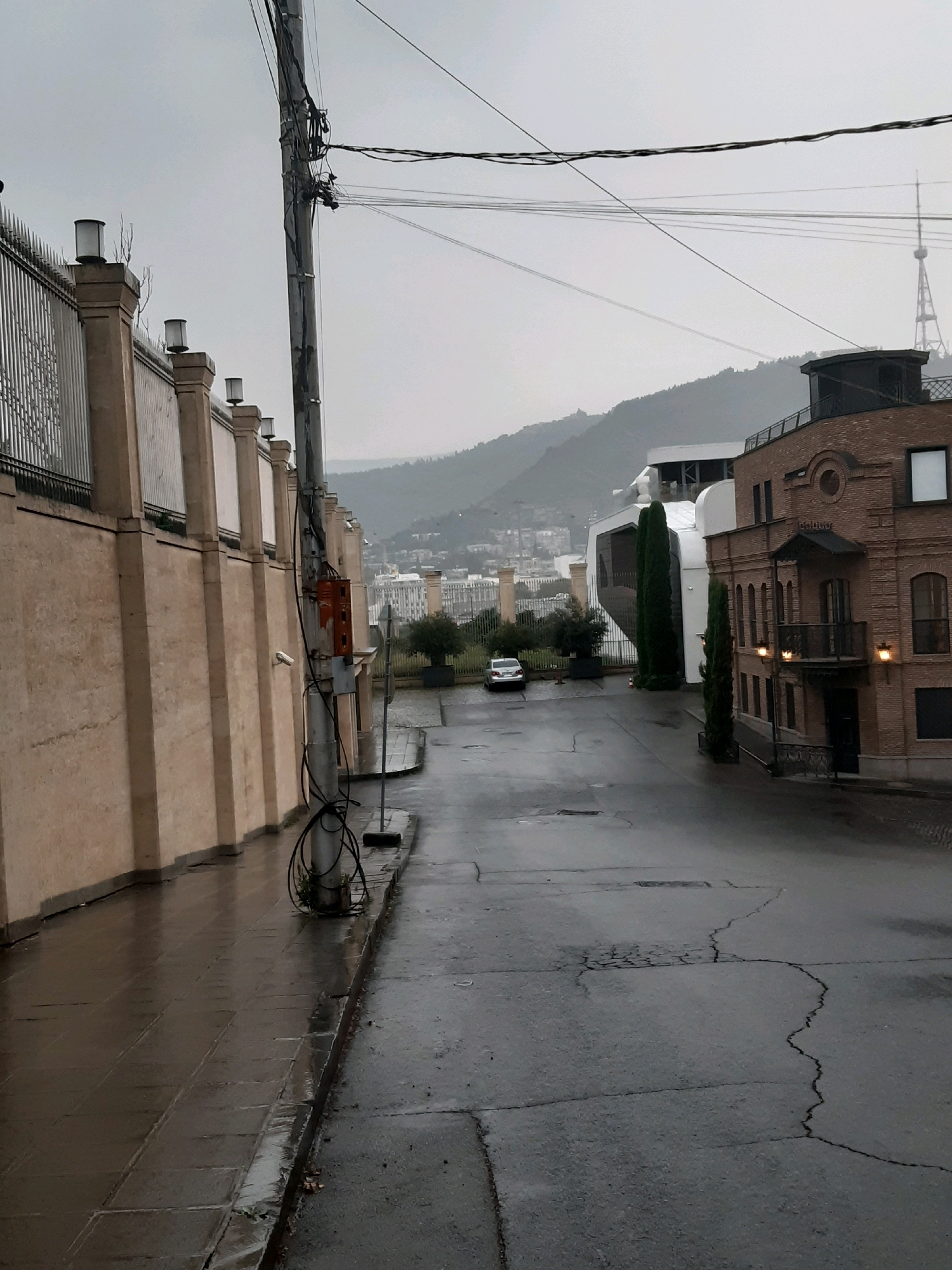 A street view of a city on an overcast day in Tbilisi, Georgia. The street is wet, indicating recent rain. On the left, a tall, beige-colored wall with decorative fencing and lights lines the sidewalk. A utility pole with numerous wires runs up the center of the image. Further down the street, on the right, is a brown brick building with a balcony and windows. Beyond that, modern white buildings and trees are visible, with a city skyline and hills in the background. A single white car is parked on the street.