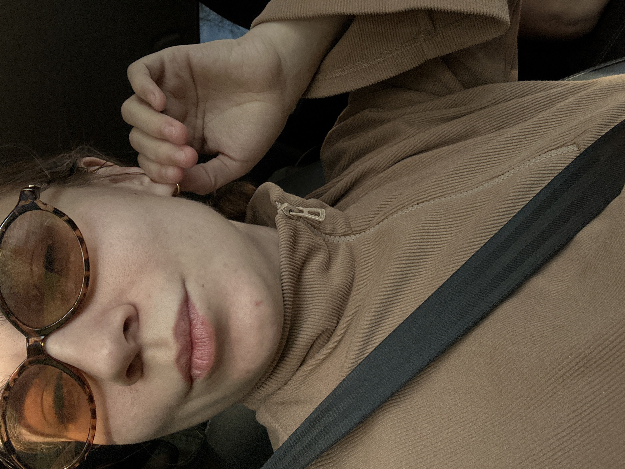 A woman is sitting in a car, wearing sunglasses and a brown corduroy turtleneck. She is wearing a seatbelt. Her hand is near her ear, which has a gold hoop earring. The location is not discernible from the image.